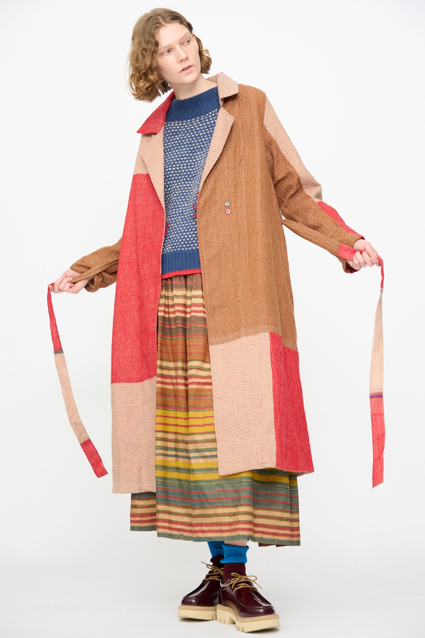 A person wearing the Injiri Qasab Wool and Silk Jacket in Red and Brown Multi over a blue sweater and a striped skirt, holding the coat ties while standing against a plain white background. This elegant piece by Injiri is crafted in India.