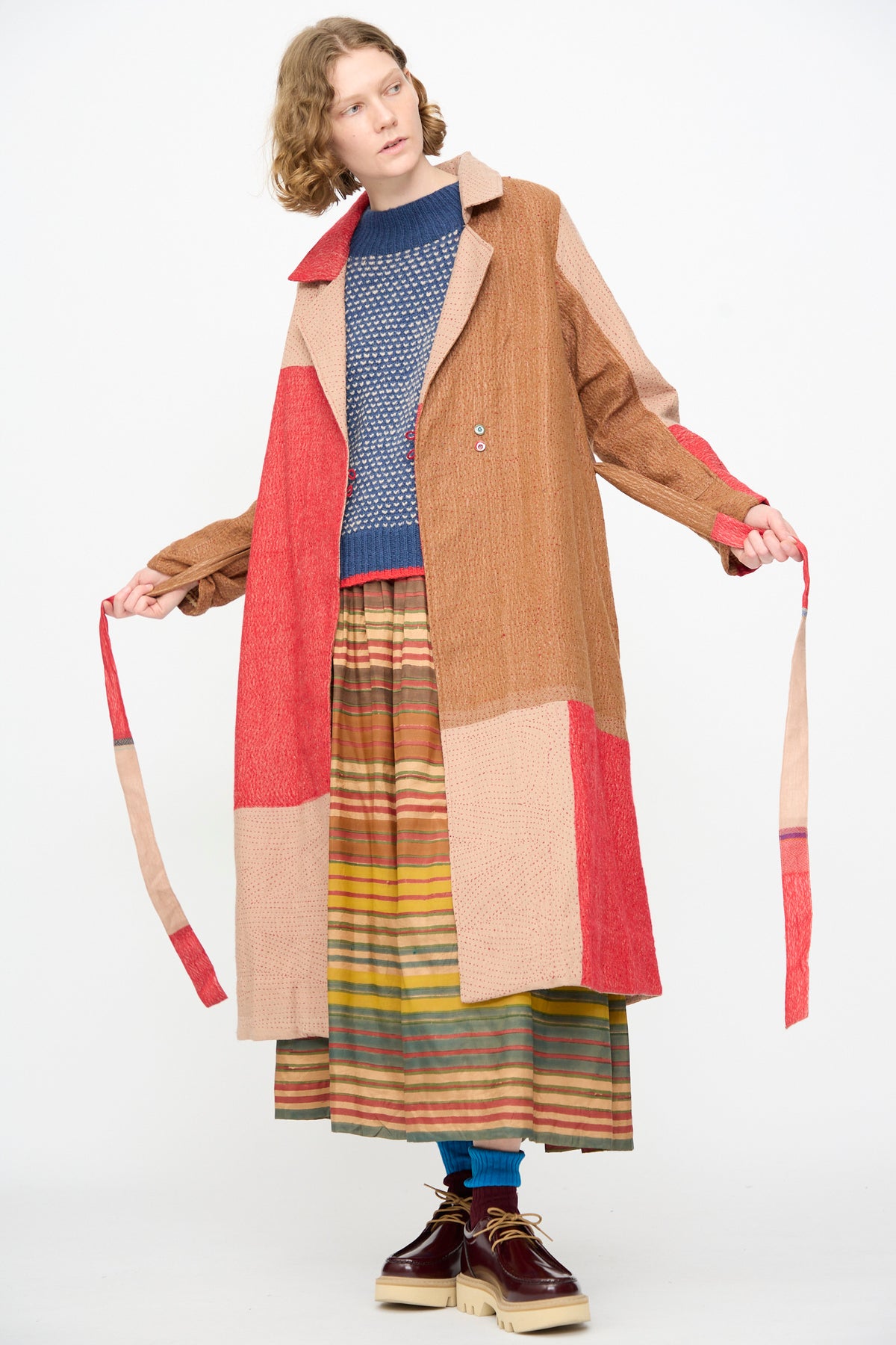 A person wearing the Injiri Qasab Wool and Silk Jacket in Red and Brown Multi over a blue sweater and a striped skirt, holding the coat ties while standing against a plain white background. This elegant piece by Injiri is crafted in India.