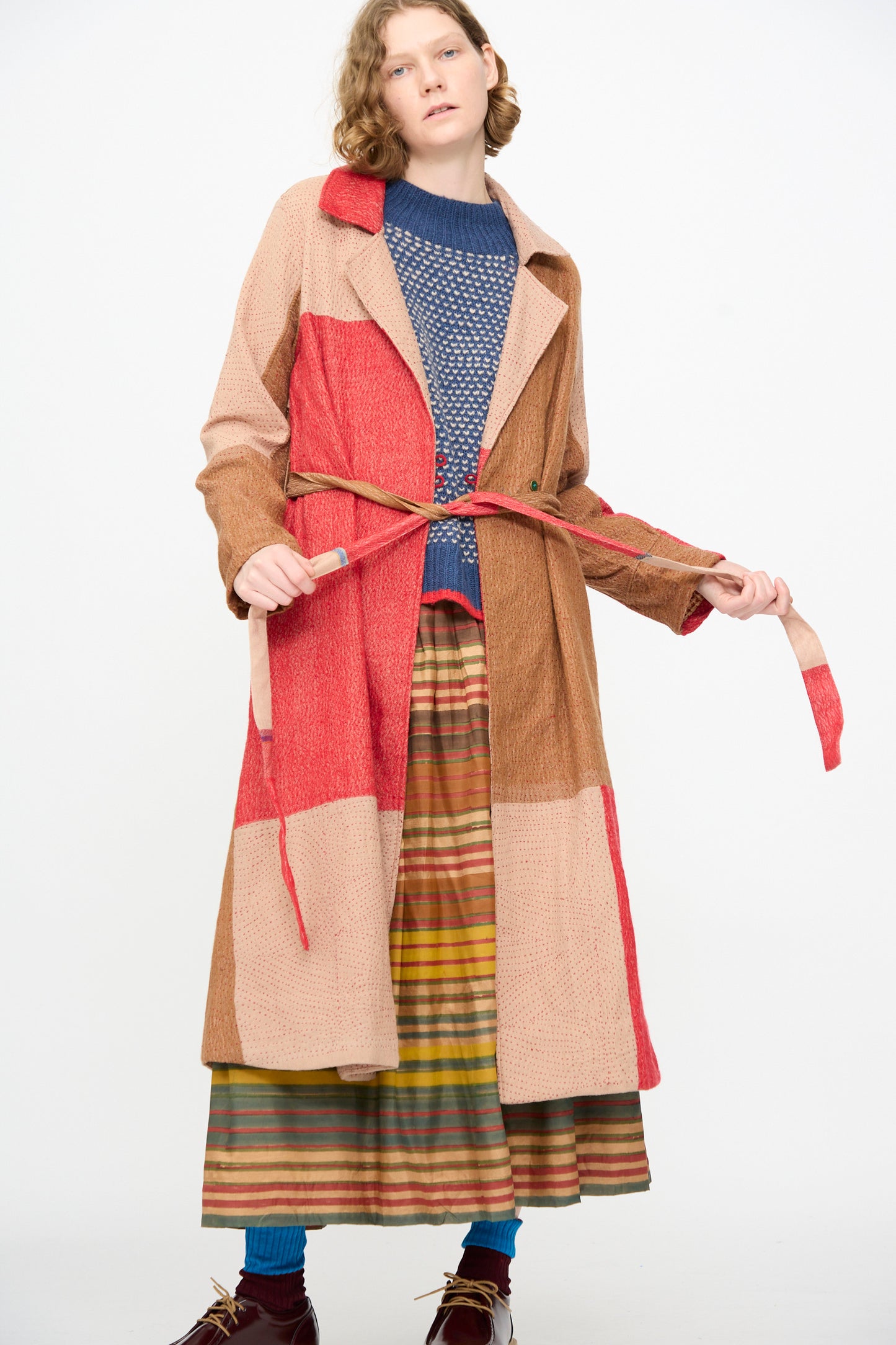 A person stands against a plain white background wearing the Injiri Qasab Wool and Silk Jacket in Red and Brown Multi, an artisanal patchwork coat crafted in India from a luxurious wool-silk blend. The ensemble is completed with a blue knit sweater, striped skirt, teal socks, and brown shoes, highlighting exceptional craftsmanship.