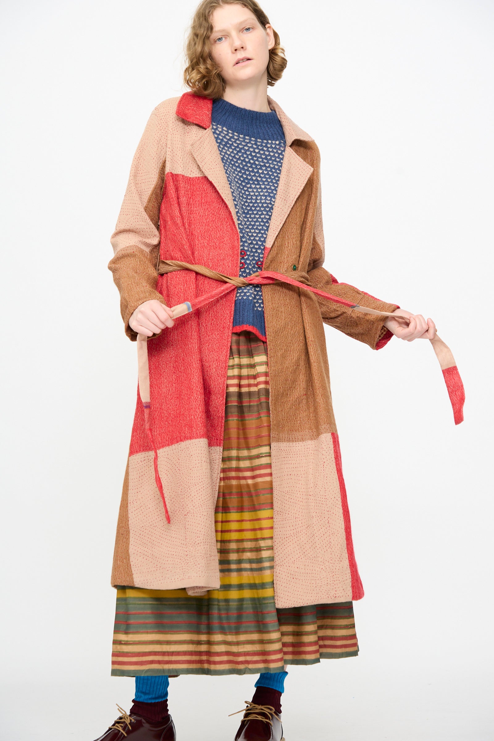A person stands against a plain white background wearing the Injiri Qasab Wool and Silk Jacket in Red and Brown Multi, an artisanal patchwork coat crafted in India from a luxurious wool-silk blend. The ensemble is completed with a blue knit sweater, striped skirt, teal socks, and brown shoes, highlighting exceptional craftsmanship.