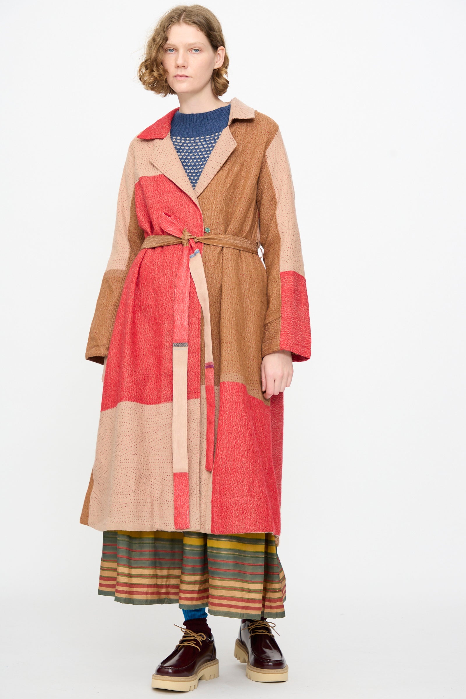 A person wearing the Injiri Qasab Wool and Silk Jacket in Red and Brown Multi—a patchwork-style, long belted coat crafted from a fine wool-silk blend—paired with a blue sweater, striped skirt, and brown shoes, stands against a plain white background.