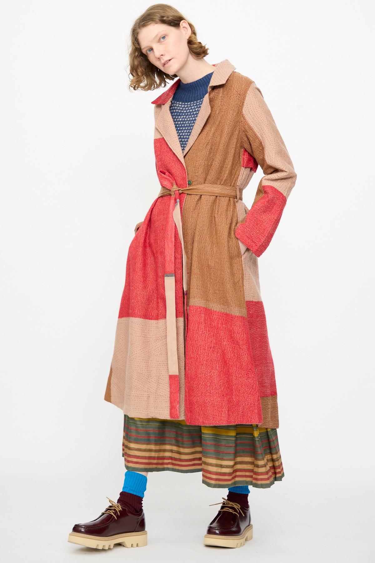 A person wearing the Injiri Qasab Wool and Silk Jacket in Red and Brown Multi—a textured, patchwork trench coat crafted from a luxurious wool-silk blend—paired with a blue sweater, striped skirt, blue socks, and brown shoes, standing against a plain white background.