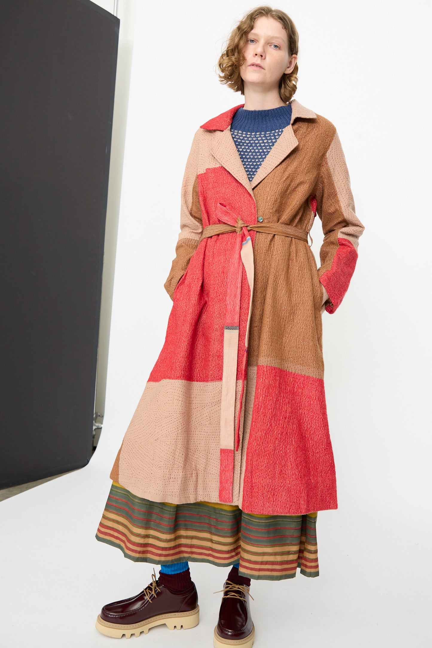 A person stands in a studio setting wearing the Injiri Qasab Wool and Silk Jacket in Red and Brown Multi—a textured long coat crafted in India from a wool-silk blend featuring red and beige color blocks—layered over a striped skirt, paired with blue socks and brown platform shoes.