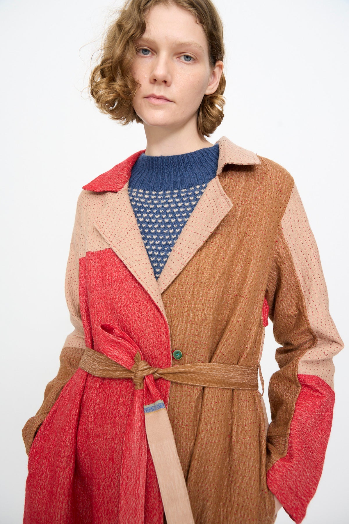 A woman with short, wavy hair wears the Injiri Qasab Wool and Silk Jacket in Red and Brown Multi—a textured, color-blocked coat crafted from a luxurious wool-silk blend and belted at the waist—layered over a blue knit sweater. She stands against a plain white background.
