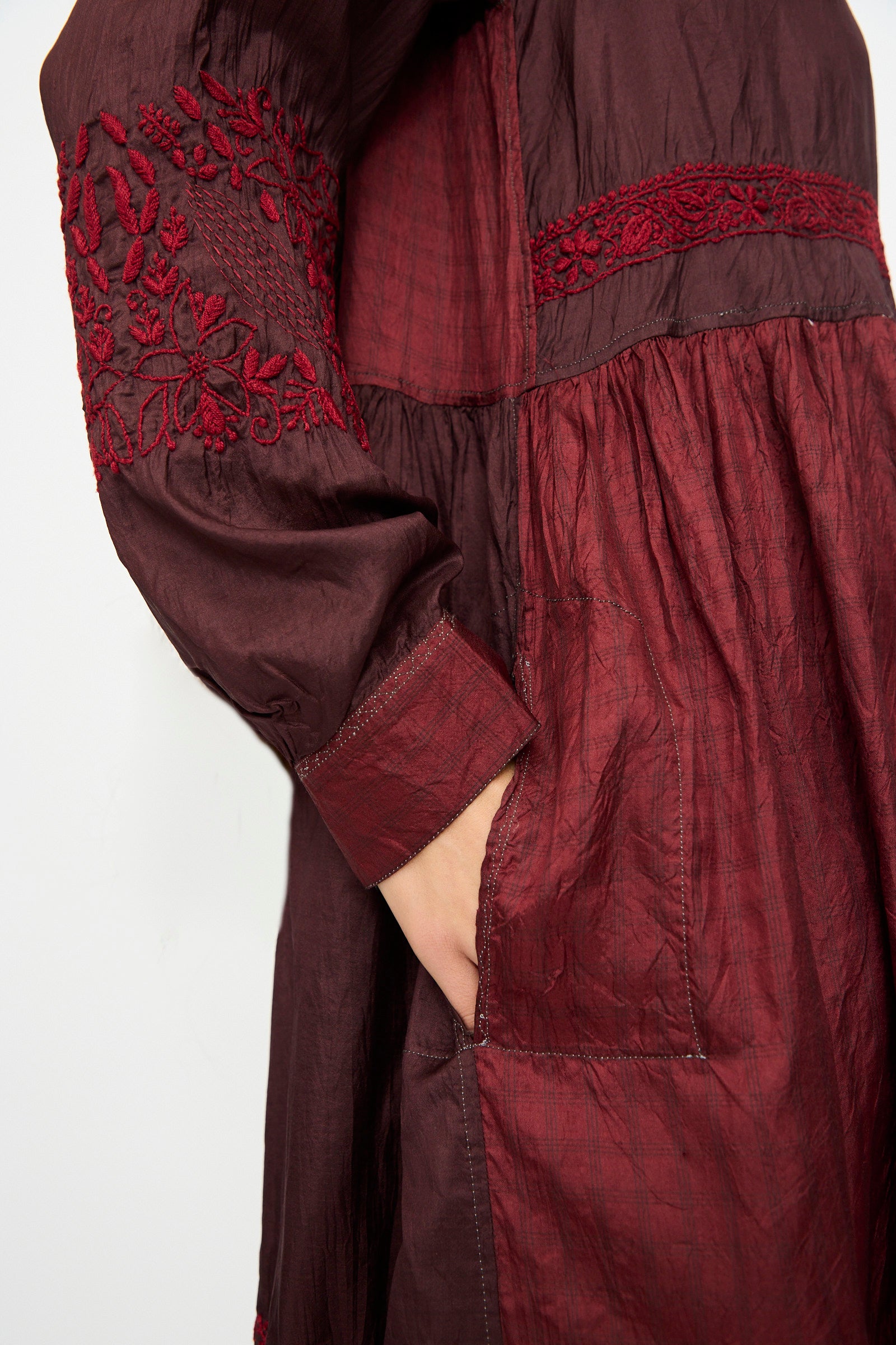 A person wearing the Silk and Cotton Dress in Red by Injiri, featuring floral embroidery on the long sleeves and a hand resting in a side pocket.