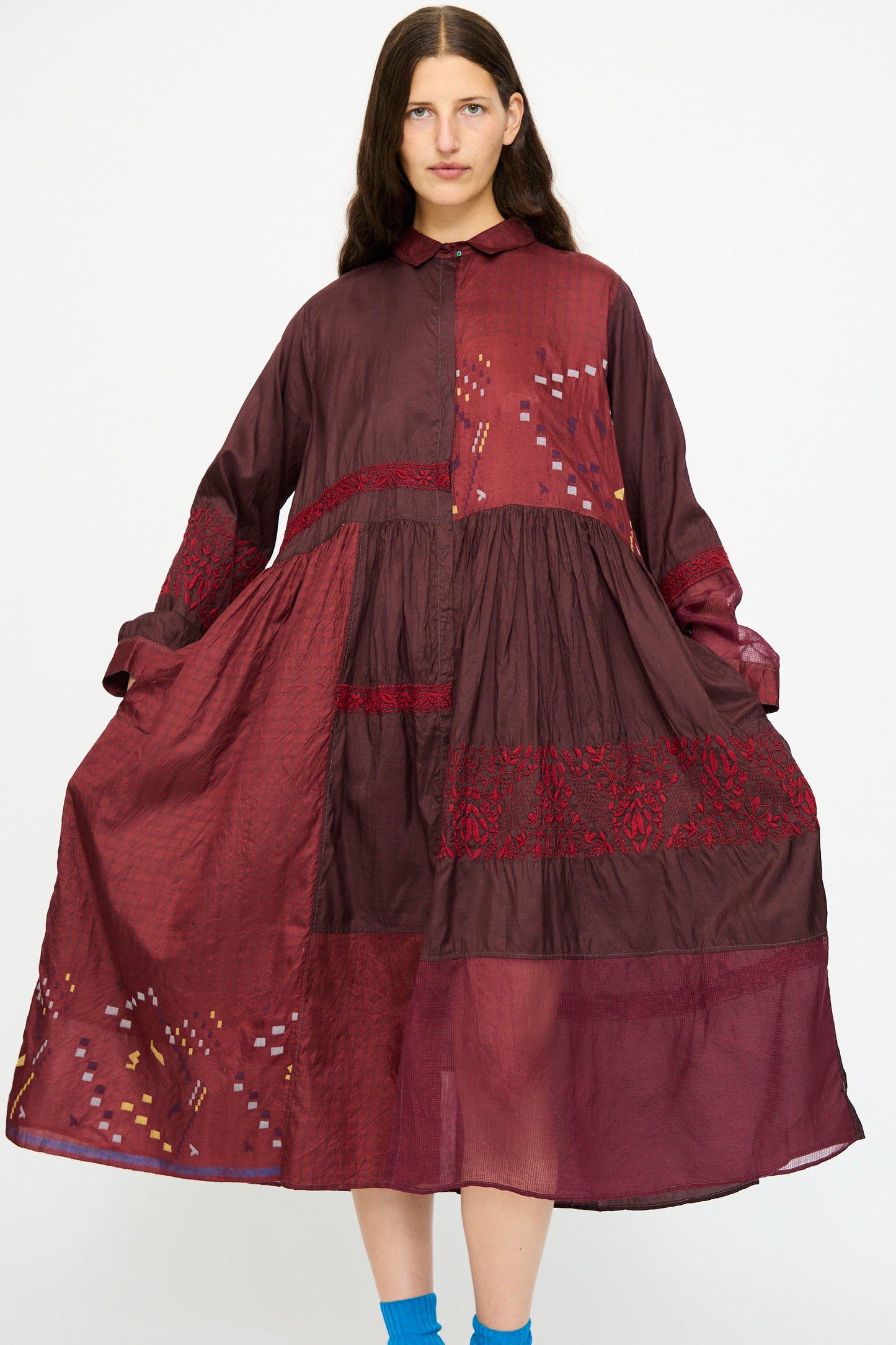 A person wears the Injiri Silk and Cotton Dress in Red, featuring loose-fitting long sleeves with embroidered and patterned patchwork details, standing against a plain white background.