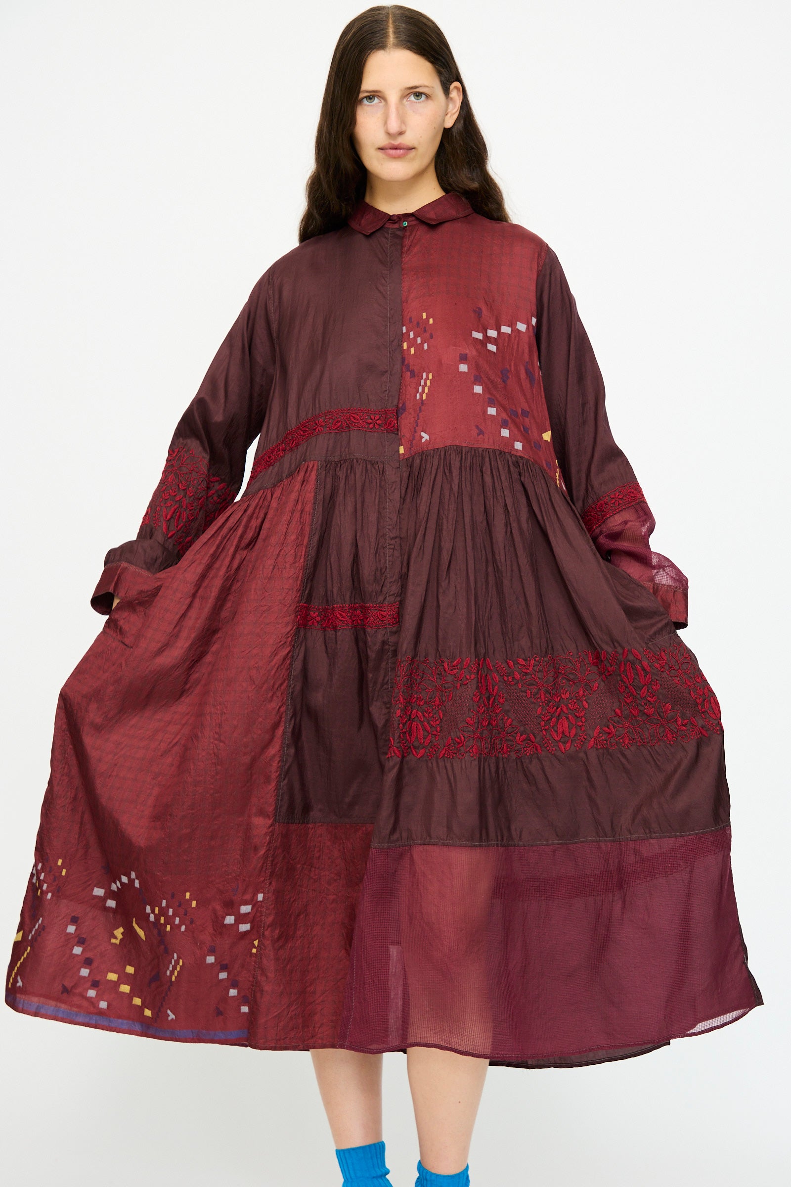 A person wears the Injiri Silk and Cotton Dress in Red, featuring loose-fitting long sleeves with embroidered and patterned patchwork details, standing against a plain white background.