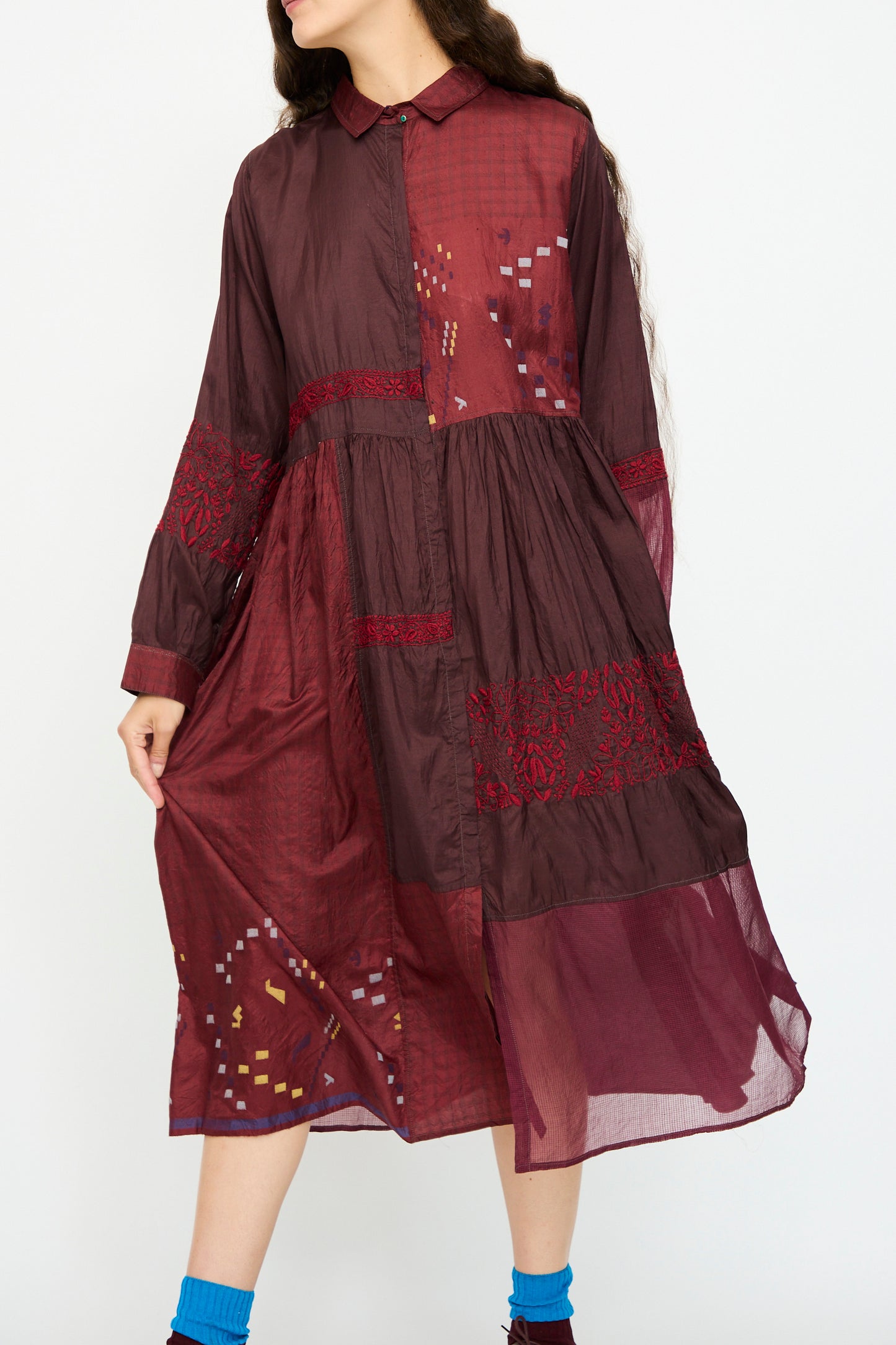 A woman wears the Injiri Silk and Cotton Dress in Red, featuring maroon and burgundy patchwork, embroidery, and sheer panels, while standing against a plain white background.