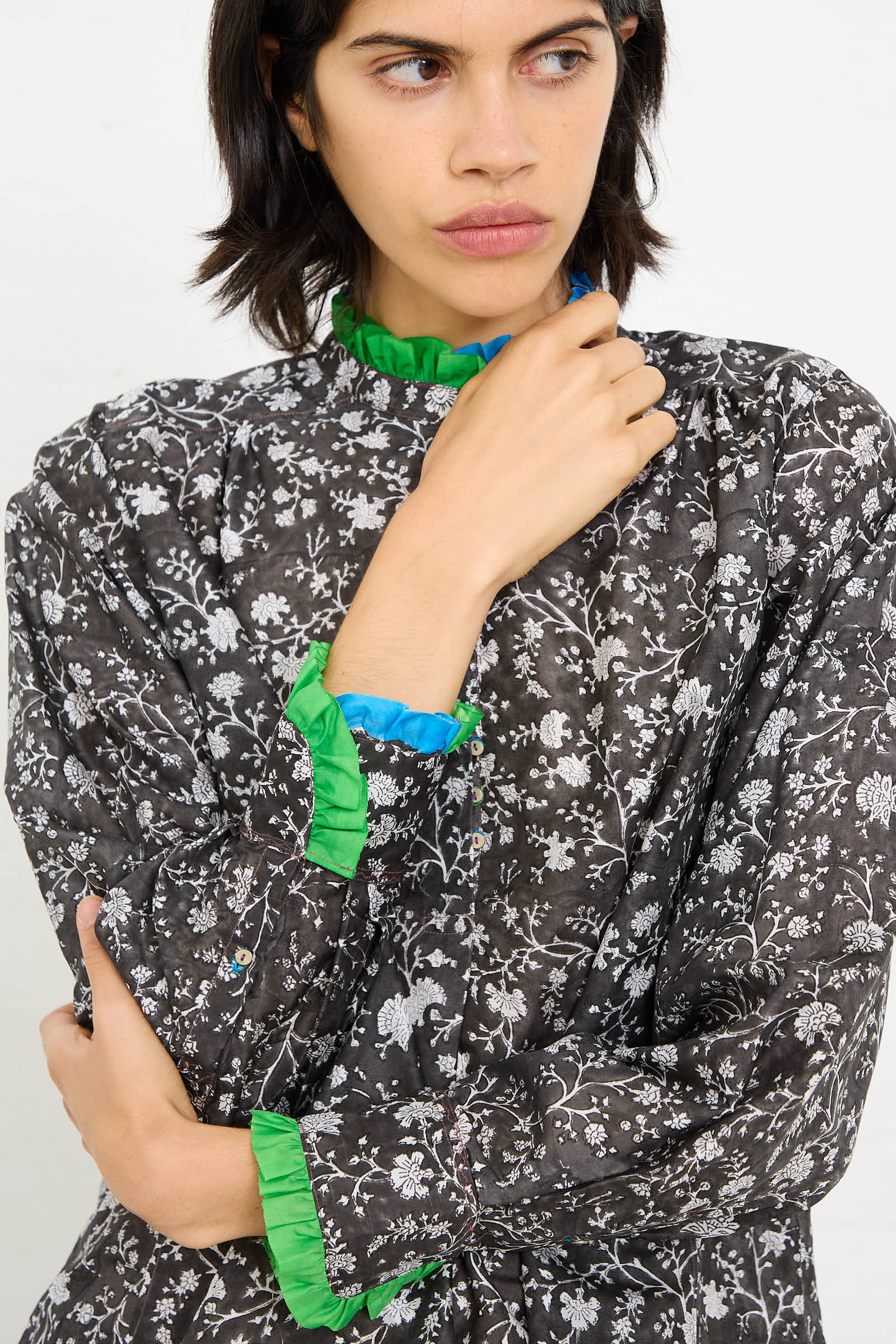 A woman with short, dark hair, wearing the Silk Blouse in Floral Print by Injiri, which features a black floral pattern with green and blue ruffled trim, holds her right hand near her chin. She gazes pensively towards the right side.