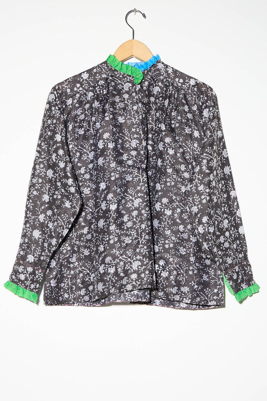 An Injiri Silk Blouse in Floral Print, with long sleeves showcasing a black and white floral pattern, green ruffles at the cuffs and collar, and a blue ruffle at the neck, is hanging on a wooden hanger.