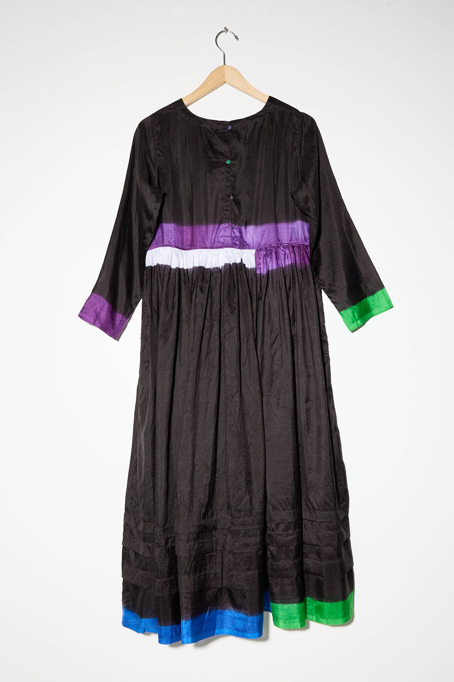 The Silk Dress in Black, Purple, and Green by Injiri hangs on a wooden hanger. This 100% silk long-sleeve dress showcases horizontal colorful stripes in purple, white, green, and blue near the chest, cuffs, and hem. It also features a tiered skirt for an added touch of elegance.