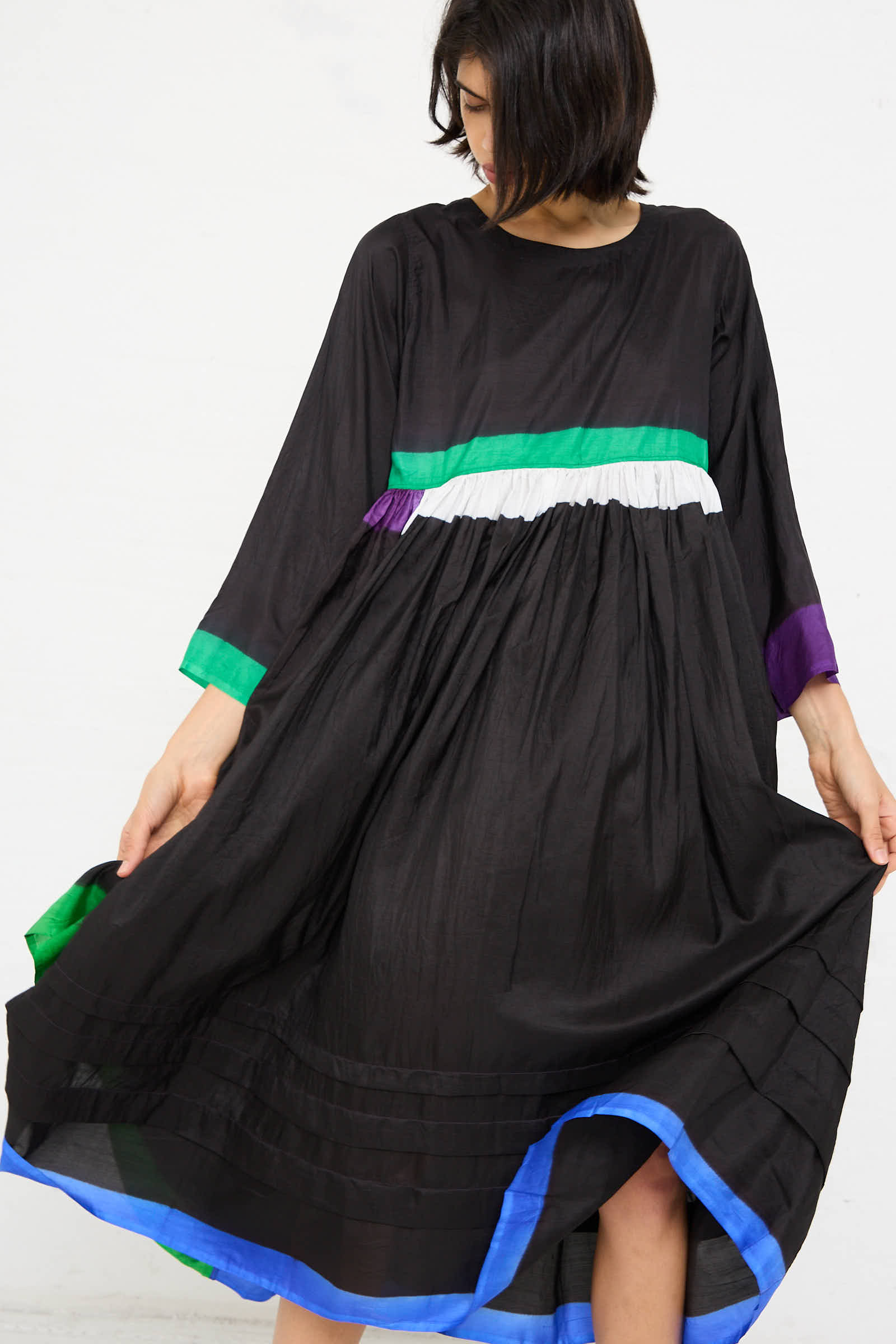 A person twirls slightly, showcasing the movement of the Silk Dress in Black, Purple and Green by Injiri. The loose-fitting dress features long sleeves and colorful accents in blue, green, purple, and white trim against a plain white background.