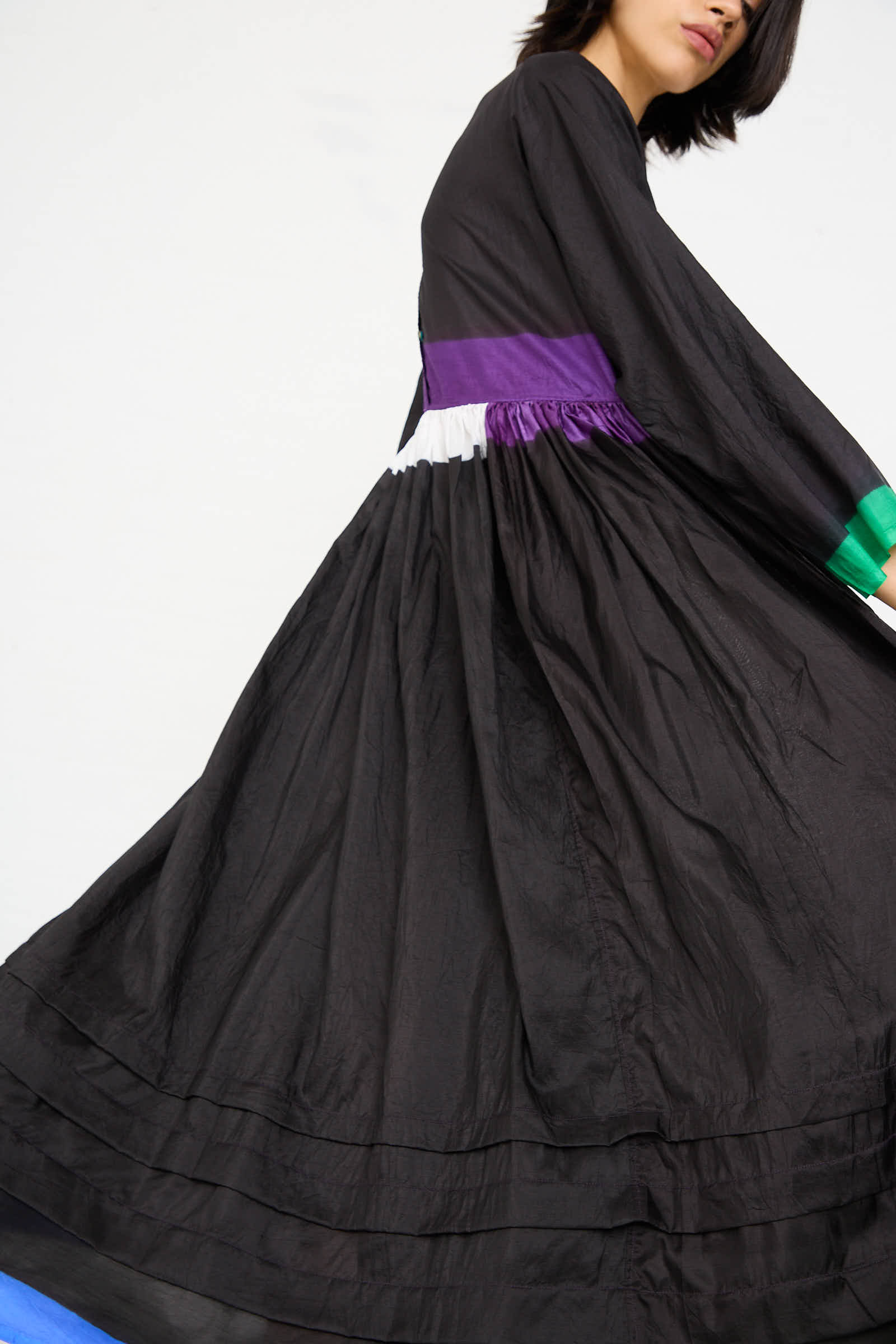 A person wearing the Injiri Silk Dress in Black, Purple and Green, characterized by its flowing long sleeves and vibrant pops of purple and white, is captured mid-motion against a plain white background.