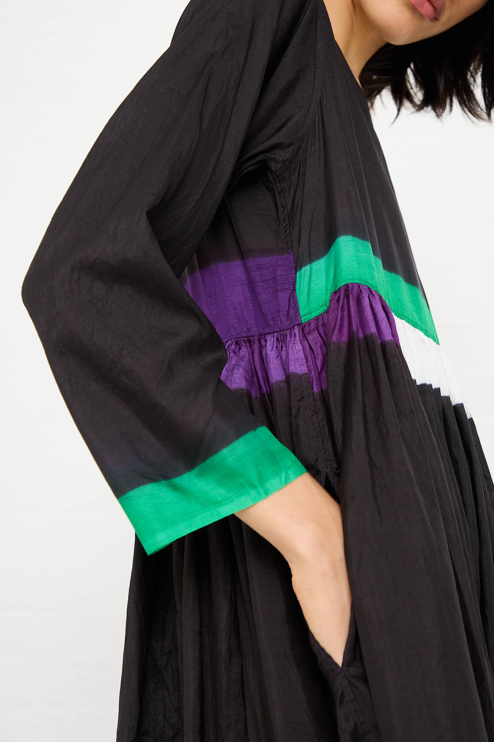 A person dressed in Injiri's Silk Dress in Black, Purple and Green, characterized by its loose-fitting long sleeves and accents of horizontal stripes with vibrant shades of green, purple, and white on the upper section. The person's hand is casually placed in the side pocket of the dress.