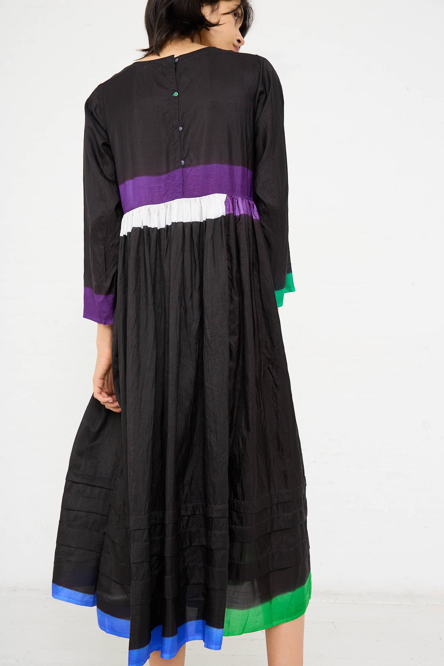 Person standing, facing away, wearing the Injiri Silk Dress in Black, Purple and Green that features colorful pops of purple, white, green, and blue horizontal stripes.