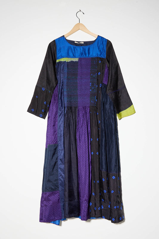 A long silk dress in shades of purple, black, and blue with a patchwork design from the brand Injiri hangs on a wooden hanger against a plain white background. The dress features accents of lime green.