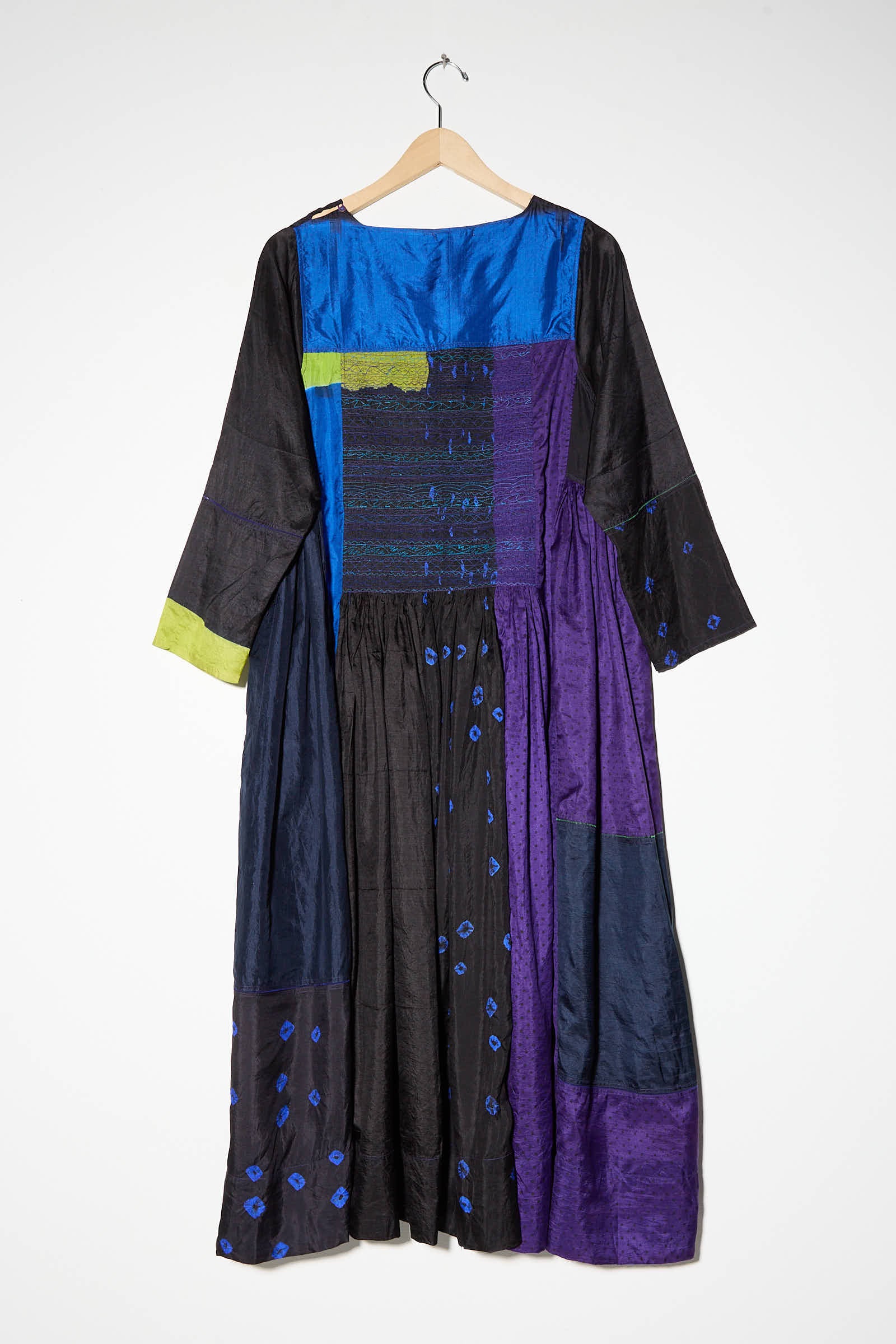 A long-sleeved Silk Dress in Purple, Black and Blue from Injiri hangs on a wooden hanger. The dress features sections of blue, purple, green, and black fabric with varying textures and patterns in a patchwork design.