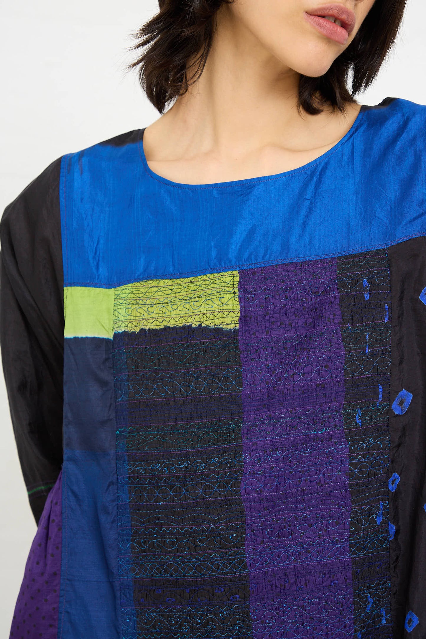 A person is wearing a Silk Dress in Purple, Black, and Blue by Injiri, featuring a multicolored patchwork design with textured patterns. The top portion of their face is not shown in the image.