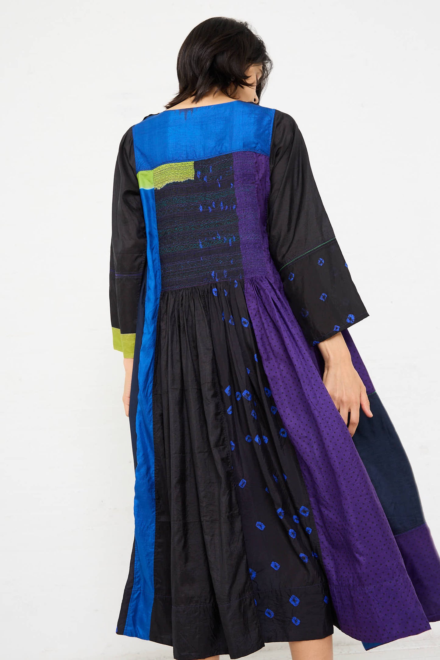 Person wearing the Silk Dress in Purple, Black, and Blue by Injiri, featuring a long-sleeve, multi-colored patchwork design with hues of blue, purple, green, and black, shown from the back.