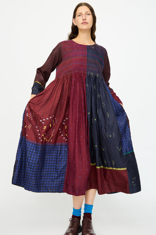 A person wears the Injiri Silk Dress in Red and Blue Multi, a loose long-sleeve style with an embroidered bodice, patterns, and textures, paired with brown shoes and blue socks.