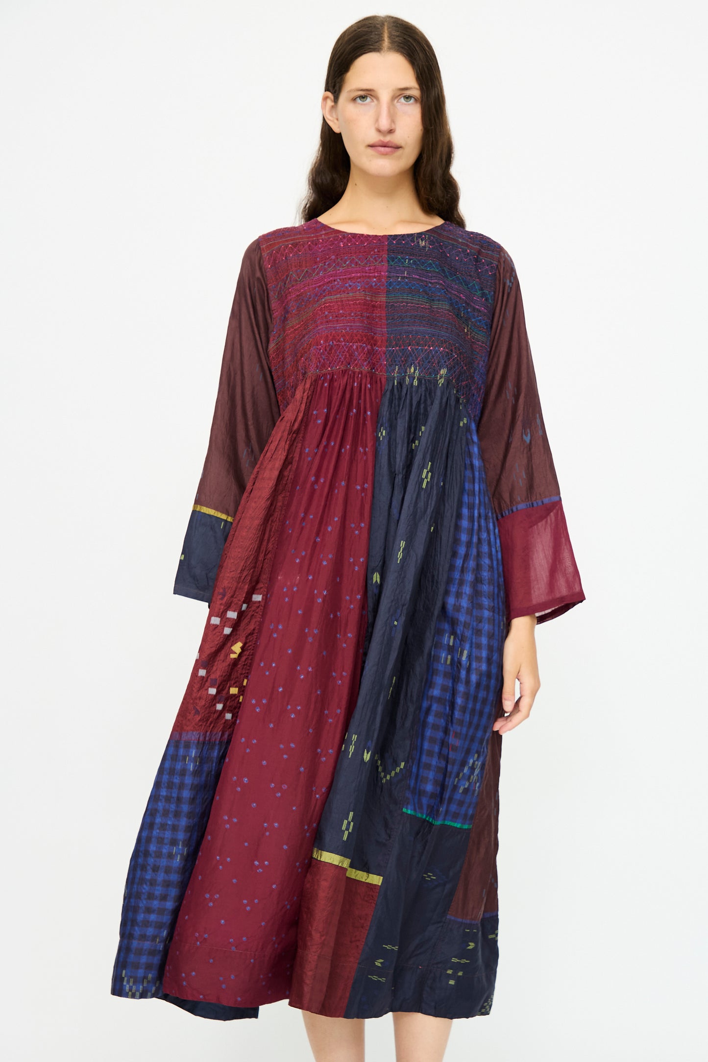 A woman stands against a plain background wearing the Injiri Silk Dress in Red and Blue Multi, a loose, long-sleeve style with patchwork patterns and an embroidered bodice.