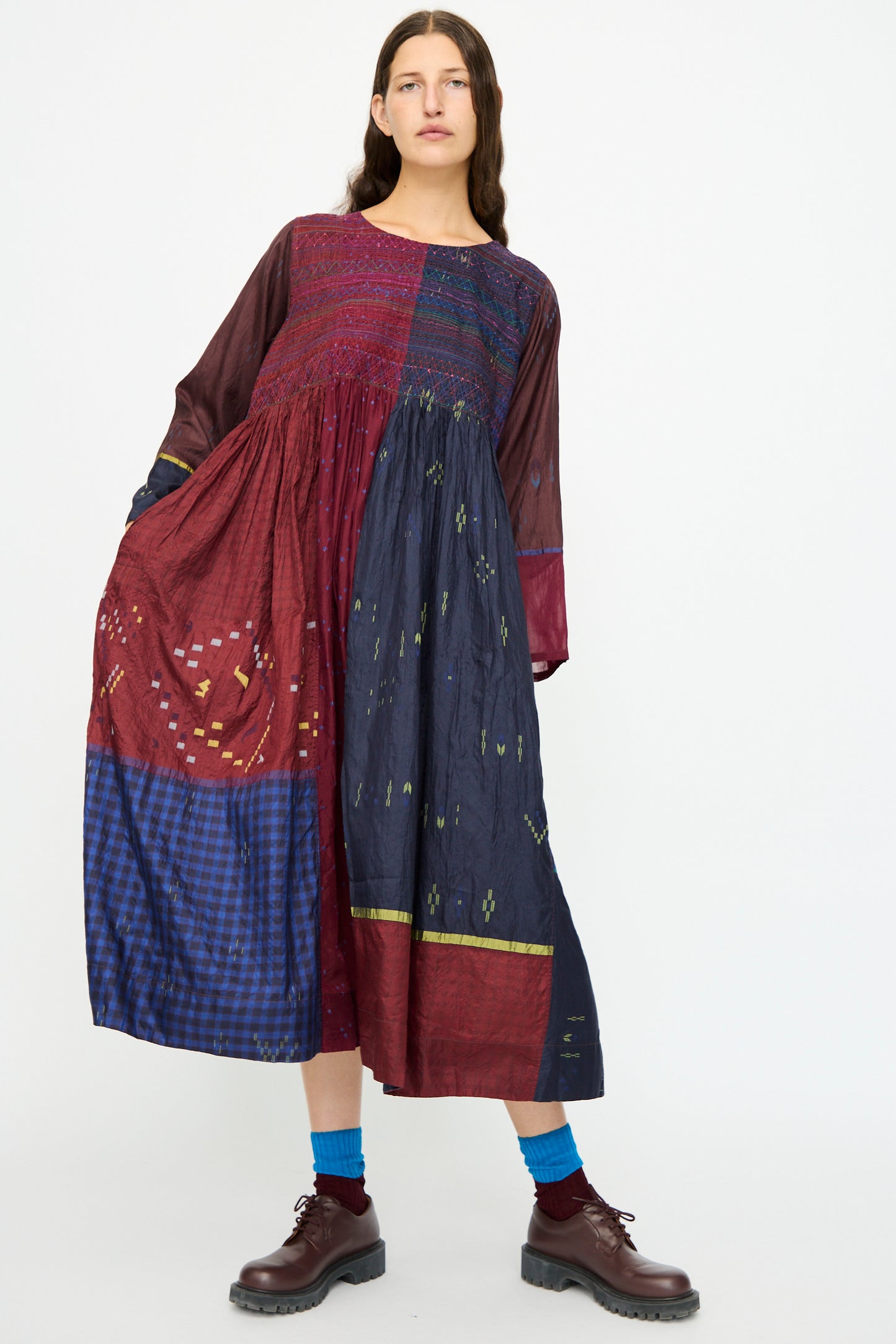 A woman stands against a plain background wearing the Injiri Silk Dress in Red and Blue Multi, featuring an embroidered bodice and color-blocked sections, paired with blue socks and brown shoes.