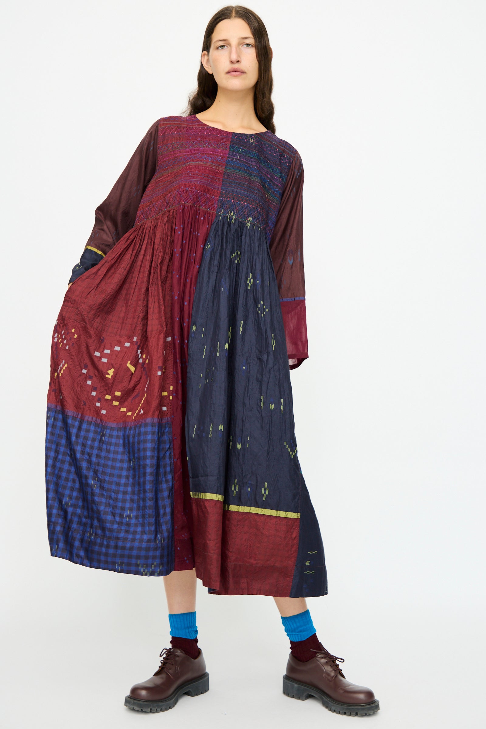 A woman stands against a plain background wearing the Injiri Silk Dress in Red and Blue Multi, featuring an embroidered bodice and color-blocked sections, paired with blue socks and brown shoes.
