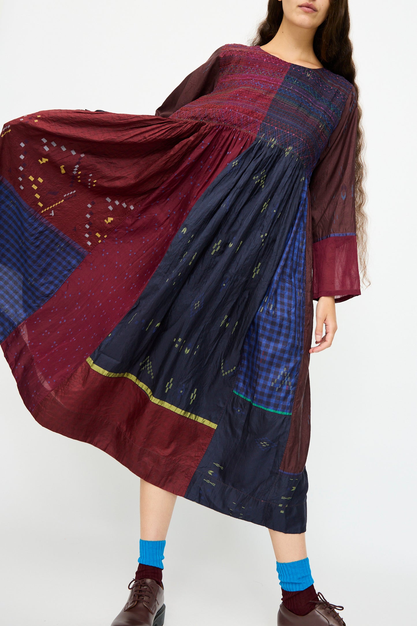 Someone wearing the Injiri Silk Dress in Red and Blue Multi, featuring long sleeves and patchwork, paired with blue socks and brown shoes. Their face is partially out of frame.