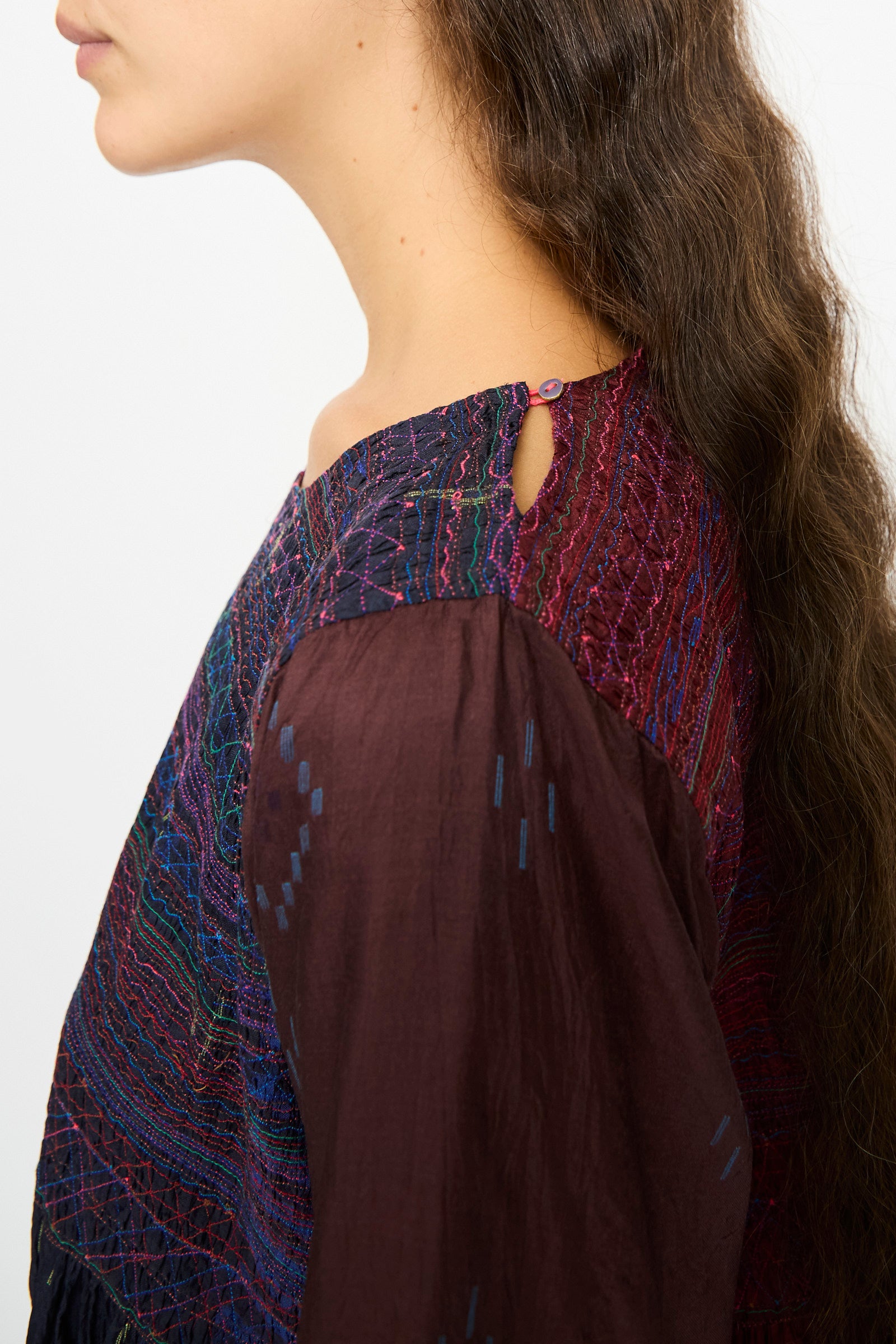 A woman with long, wavy brown hair is shown from the side wearing the Injiri Silk Dress in Red and Blue Multi, featuring an embroidered bodice and button detail at the neckline.