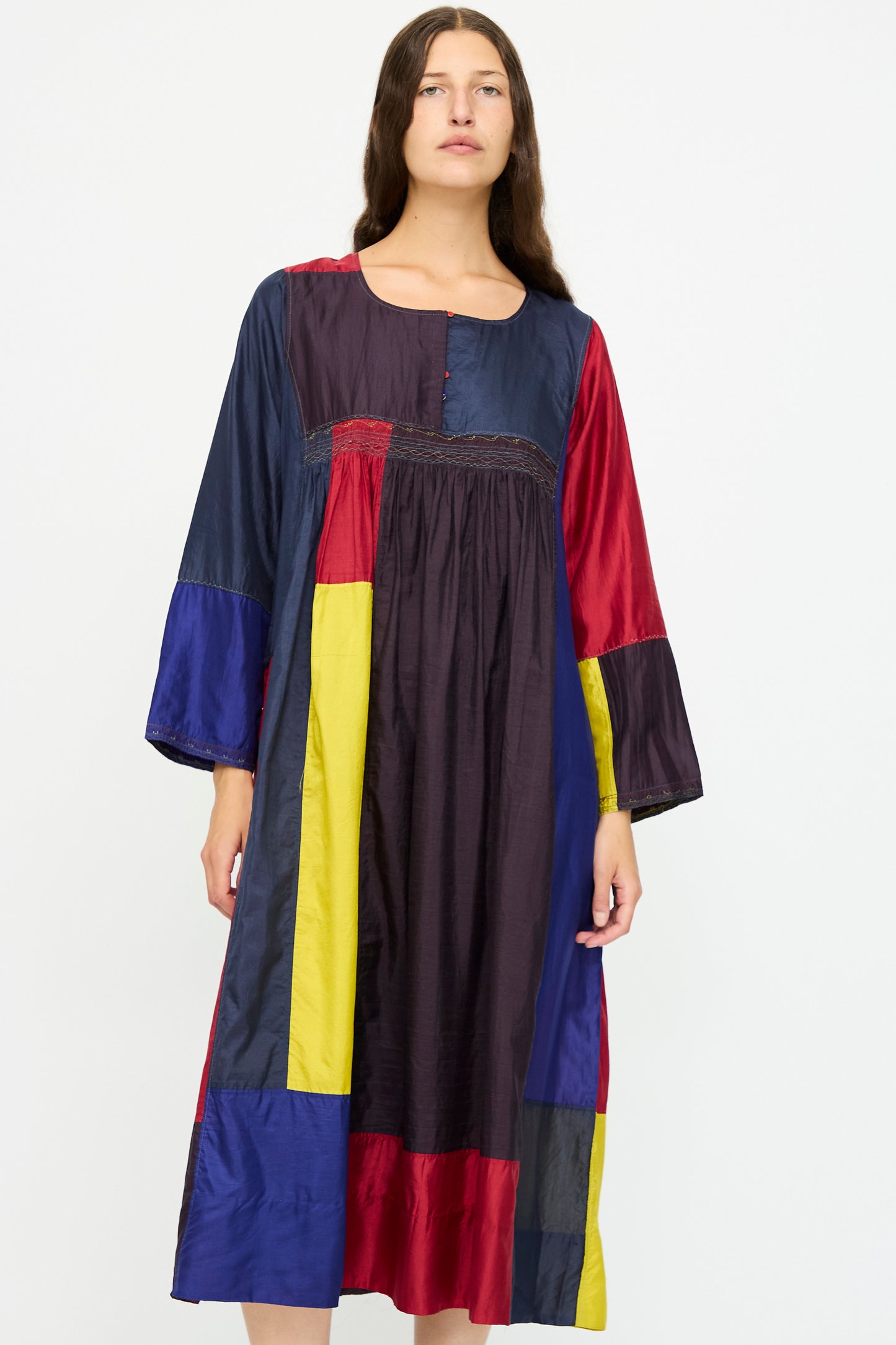 A person wears the Injiri Silk Dress in Red, Blue and Yellow Multi—a relaxed fit, long-sleeve dress with color-block panels—while standing against a plain white background.