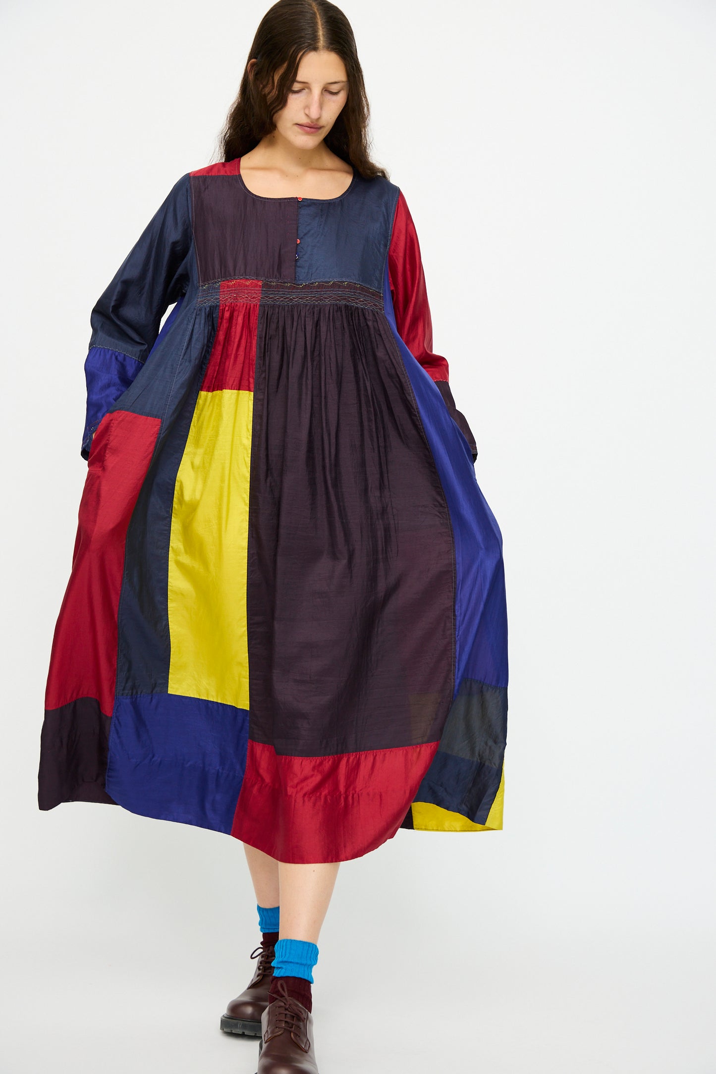 A woman wears the Injiri Silk Dress in Red, Blue and Yellow Multi, featuring a relaxed fit and long sleeves, paired with blue socks and brown shoes.