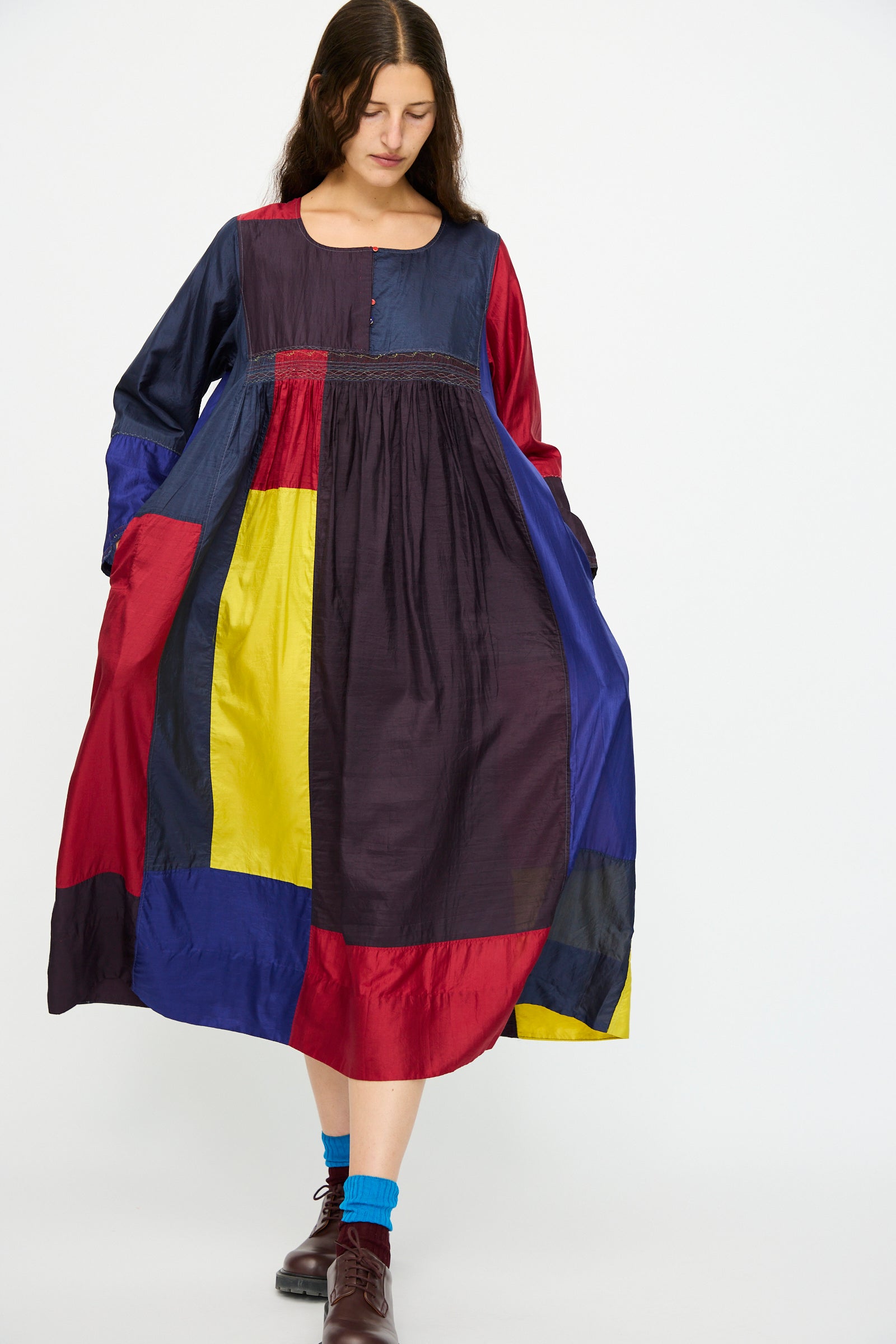 A woman wears the Injiri Silk Dress in Red, Blue and Yellow Multi, featuring a relaxed fit and long sleeves, paired with blue socks and brown shoes.