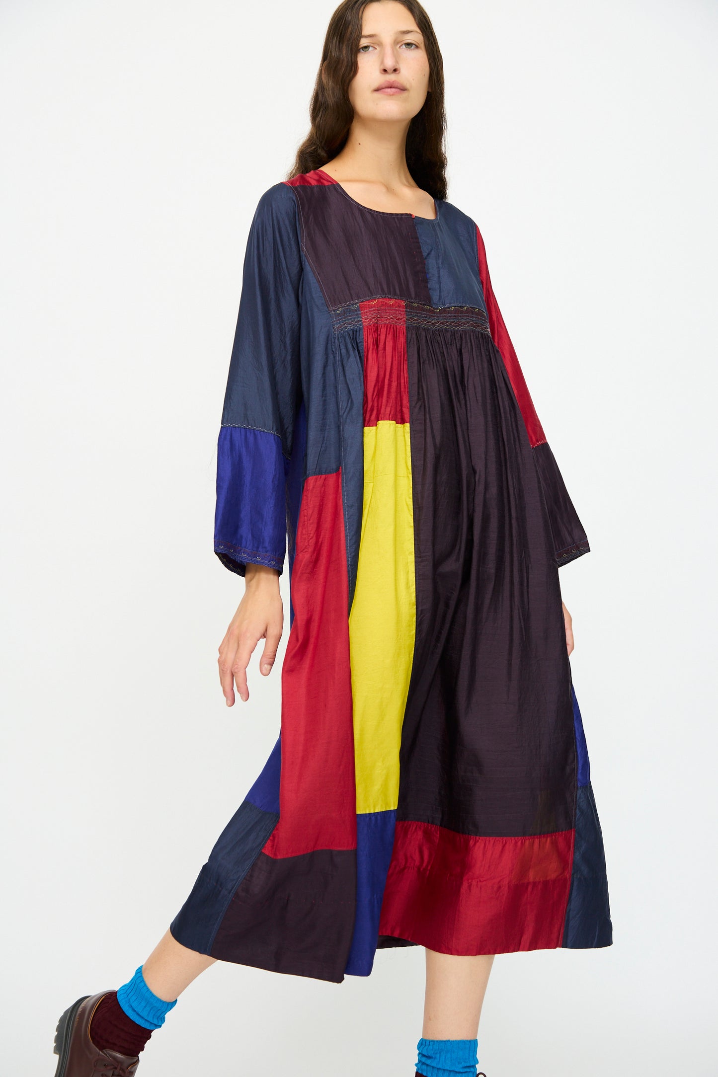 A woman wears the Injiri Silk Dress in Red, Blue and Yellow Multi, featuring a relaxed fit and long sleeves, styled with blue socks and brown shoes.