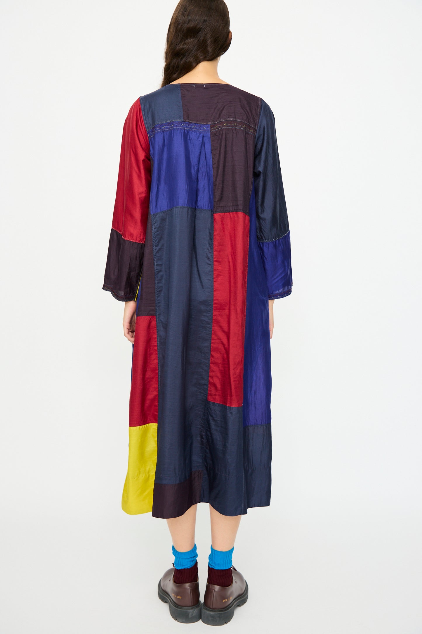 A person faces away, wearing the Injiri Silk Dress in Red, Blue and Yellow Multi—relaxed fit and long-sleeved—paired with blue socks and brown shoes.