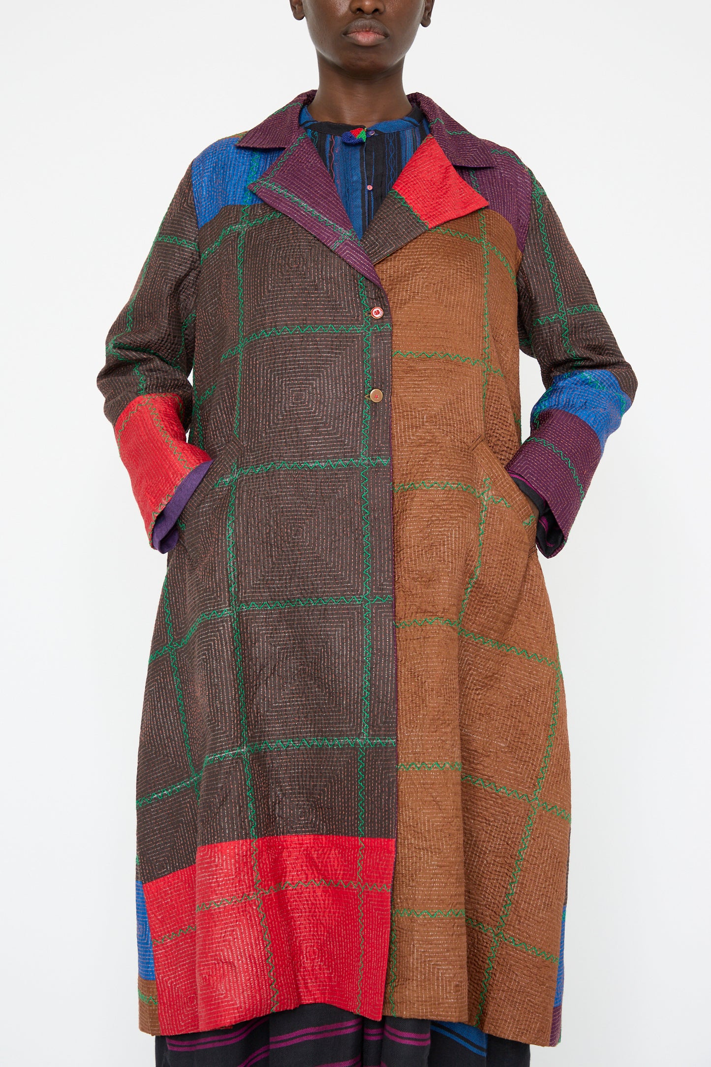 A person wearing the Silk Jacket in Brown and Multi by Injiri, a multicolored patchwork silk long coat with a plaid pattern, showcasing sections in red, brown, blue, and green.