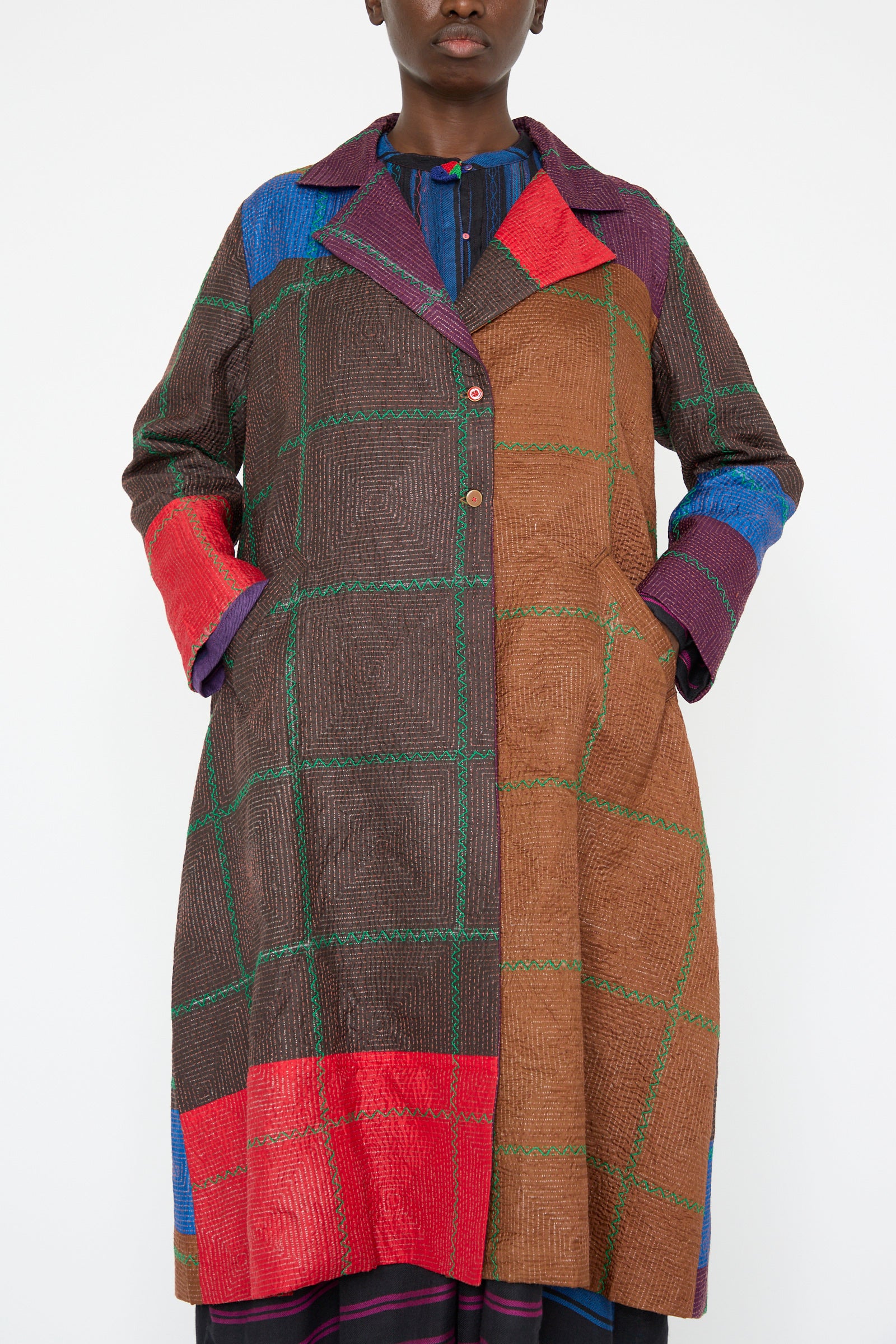 A person wearing the Silk Jacket in Brown and Multi by Injiri, a multicolored patchwork silk long coat with a plaid pattern, showcasing sections in red, brown, blue, and green.