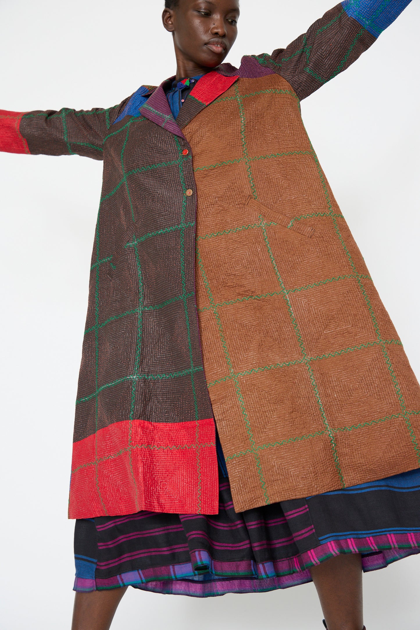 A person is wearing the Injiri Silk Jacket in Brown and Multi, featuring a grid pattern with vibrant colors of red, blue, brown, and green over a striped dress. Their arms are raised slightly against the plain white background.