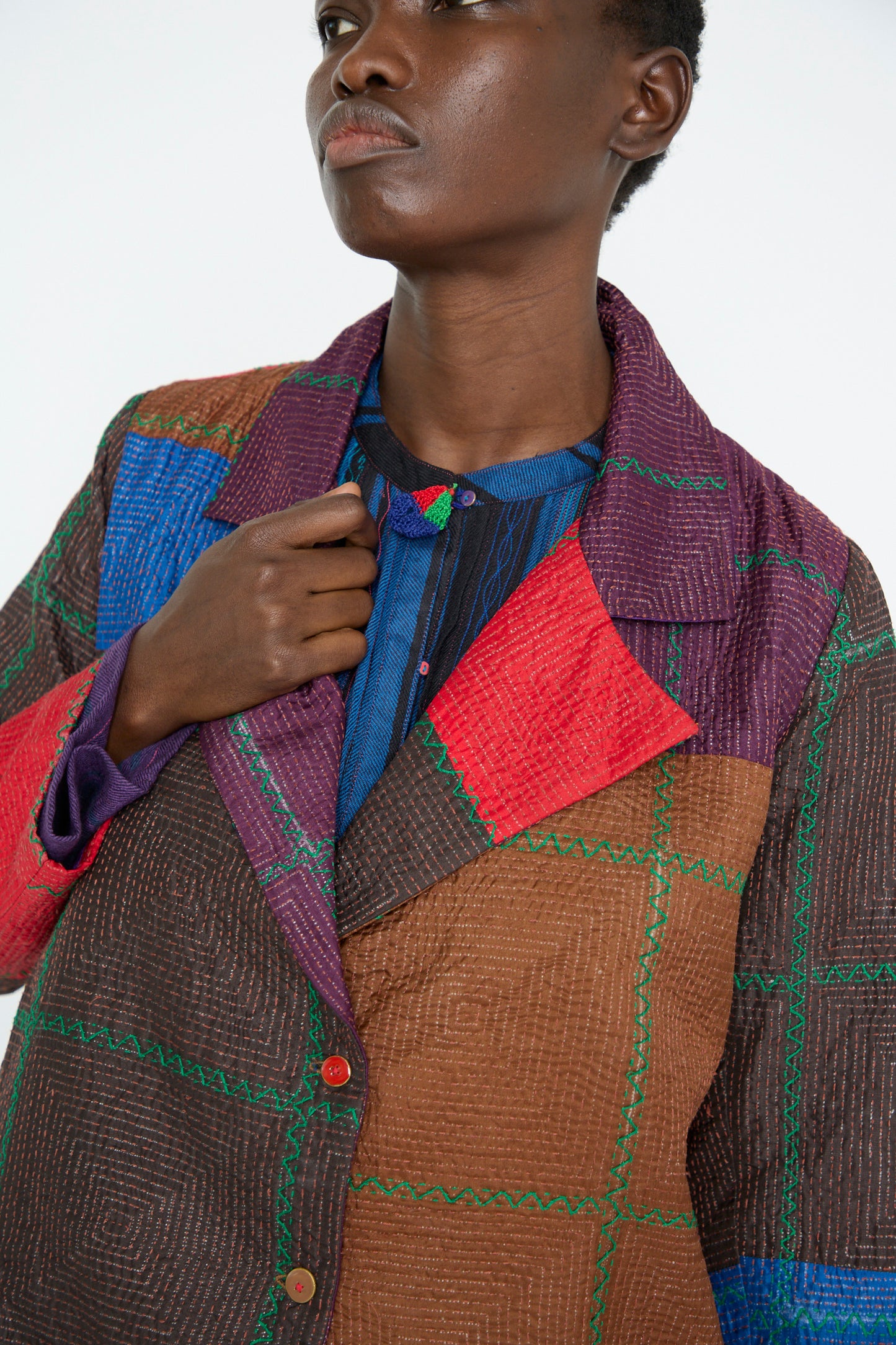 A person wearing the Injiri Silk Jacket in Brown and Multi, featuring a vibrant patchwork design with blue and red checkered patterns, holding the lapel.