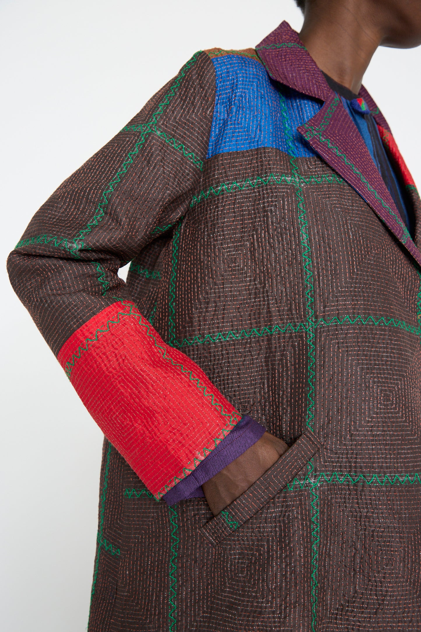 A person wearing the Silk Jacket in Brown and Multi by Injiri, featuring vibrant red, blue, and green square patterns in a patchwork design, with their hand resting in the pocket.