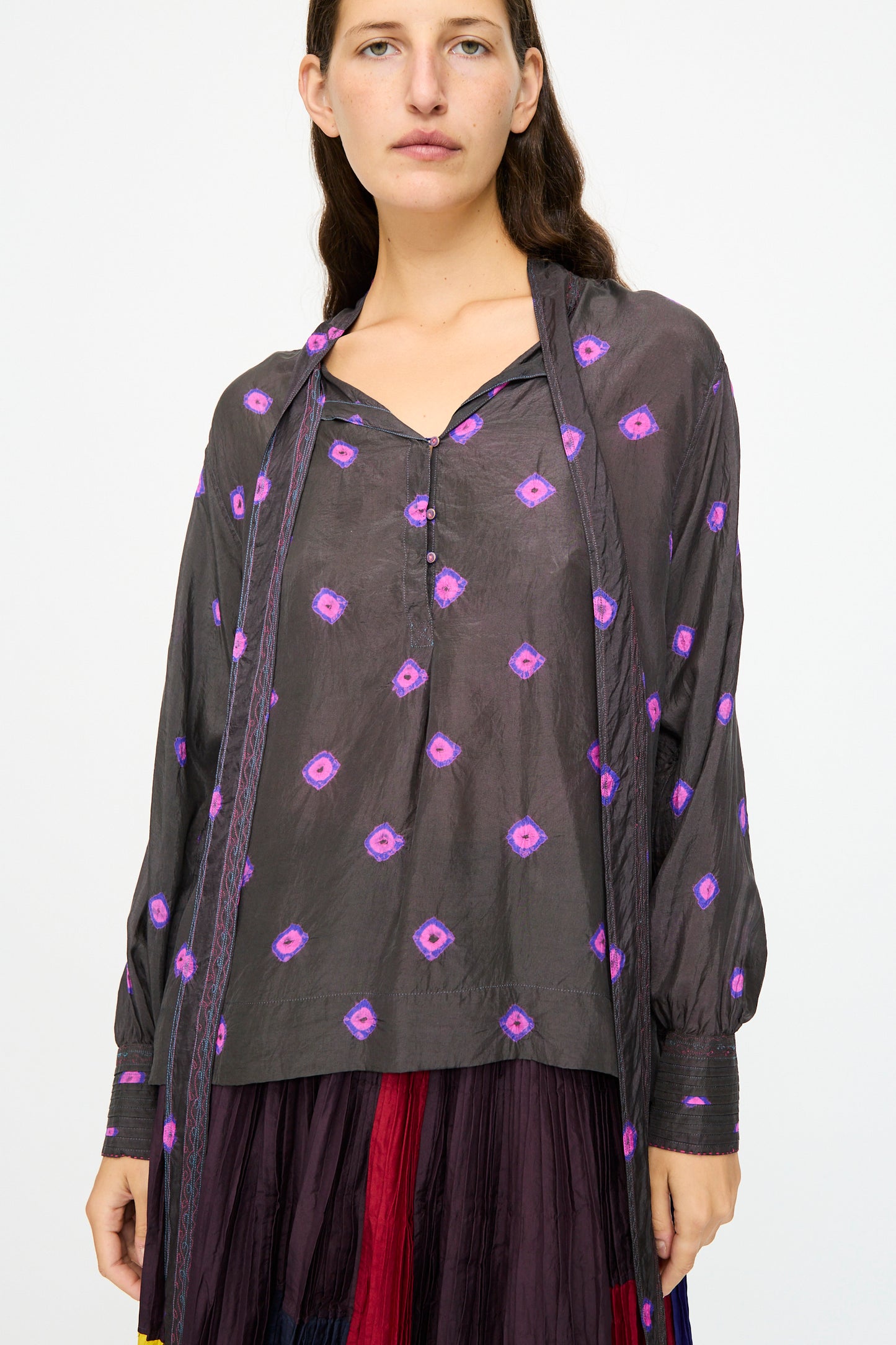 A woman wears the Injiri Silk Shirt in Purple, featuring loose long sleeves and geometric patterns, paired with a multicolored skirt against a plain light background.