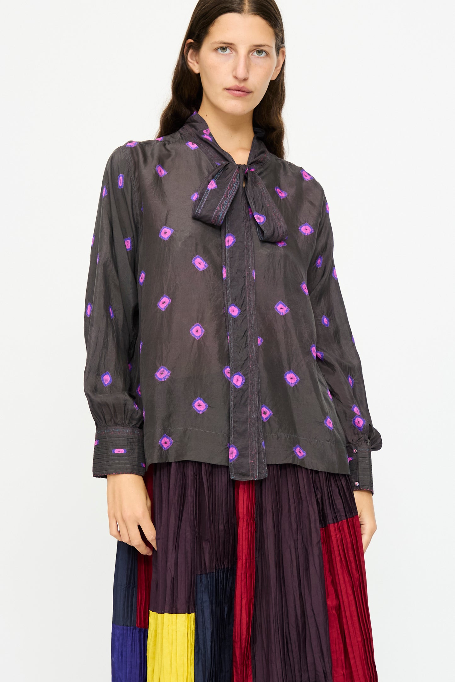A woman wears the Injiri Silk Shirt in Purple with long sleeves and a neck bow, paired with a pleated skirt featuring red, yellow, navy, and purple color blocks.