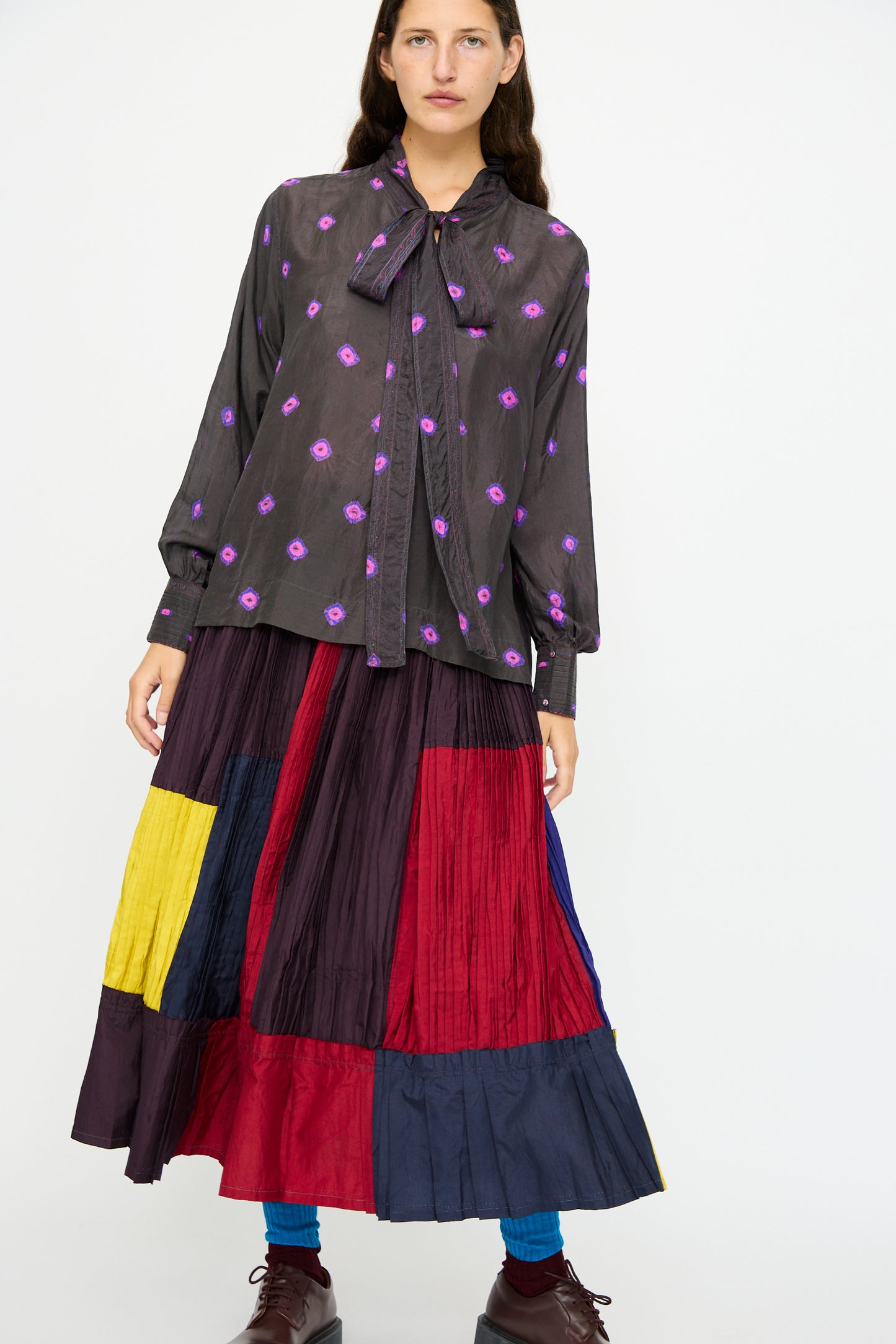 A woman wears the Injiri Silk Shirt in Purple, styled with a long, pleated skirt featuring bold color blocks of red, yellow, navy, and burgundy.