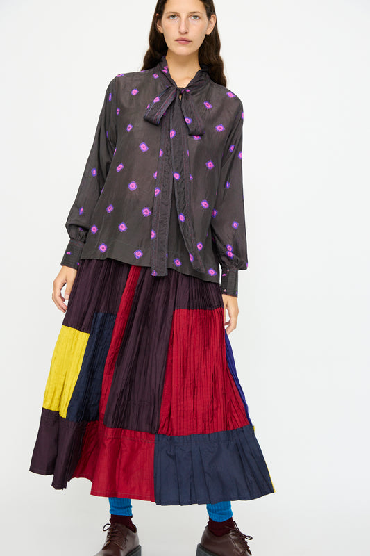 A woman wears the Injiri Silk Shirt in Purple, styled with a long, pleated skirt featuring bold color blocks of red, yellow, navy, and burgundy.