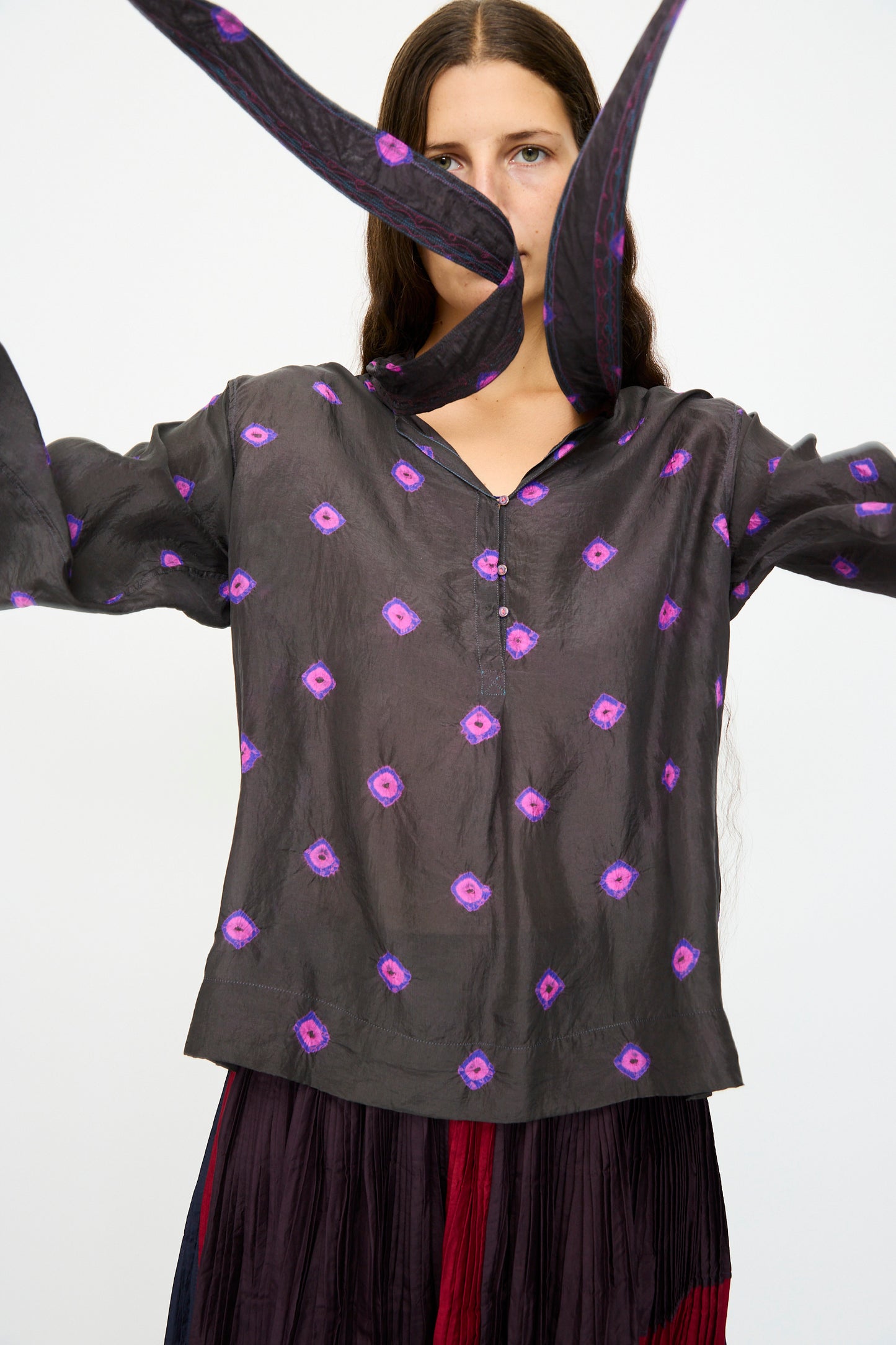 A woman in the Injiri Silk Shirt in Purple with long sleeves stands with arms raised, a matching scarf floating before her face.