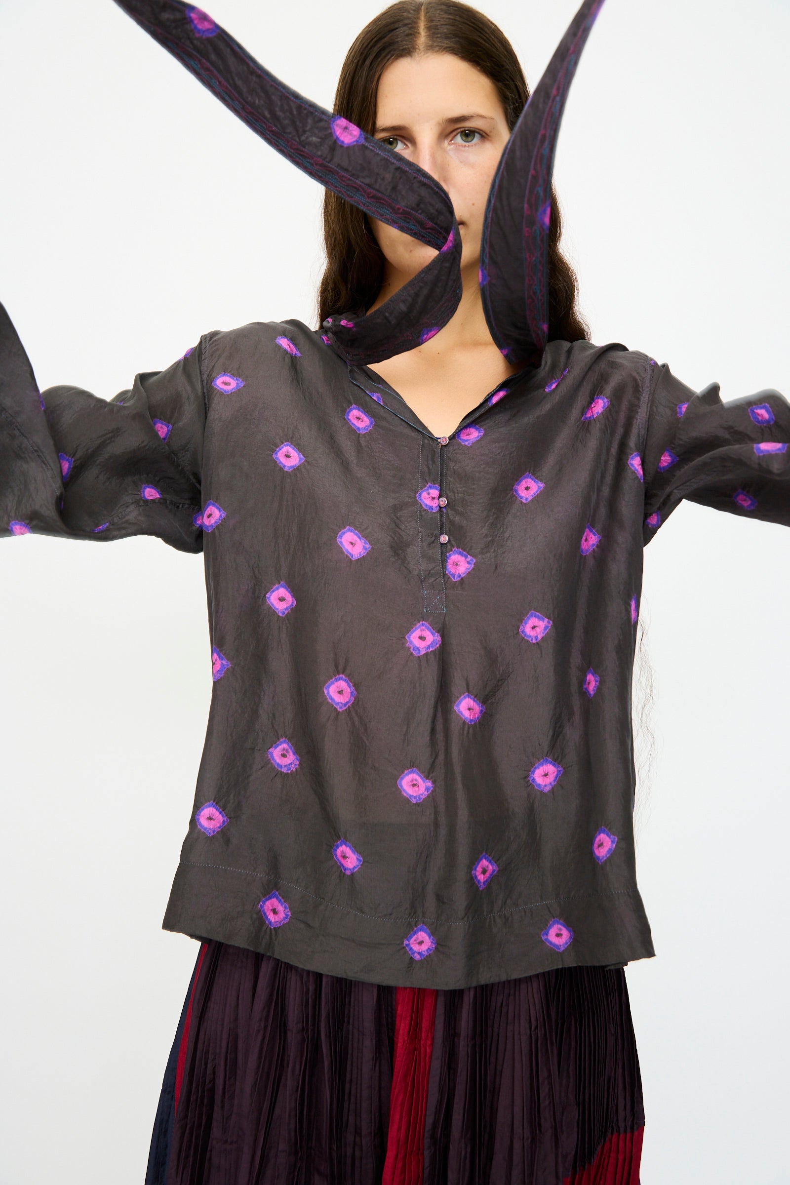 A woman in the Injiri Silk Shirt in Purple with long sleeves stands with arms raised, a matching scarf floating before her face.
