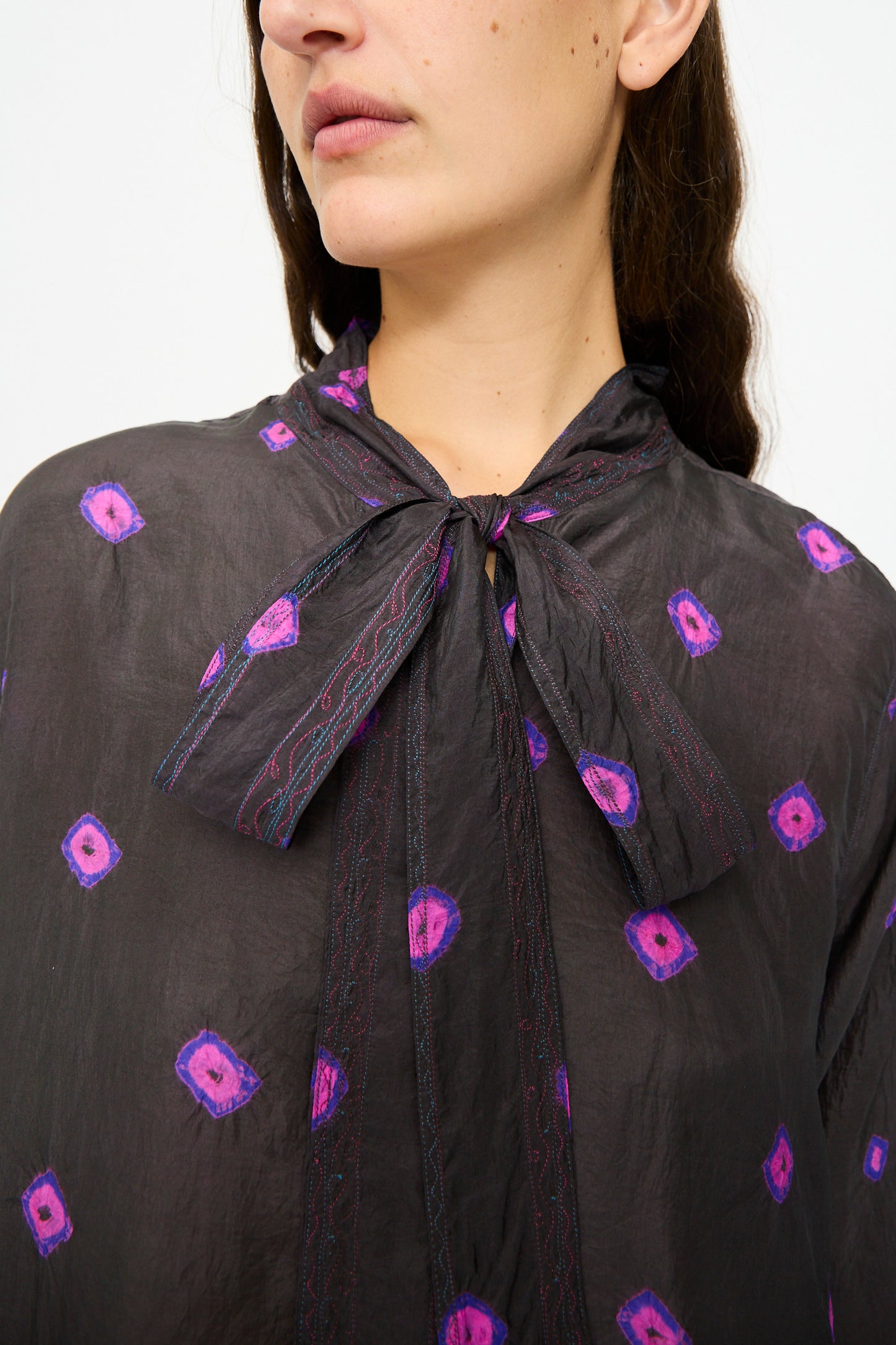 A woman wearing the Injiri Silk Shirt in Purple, a sheer black long sleeve blouse featuring purple and pink geometric patterns and a necktie bow.