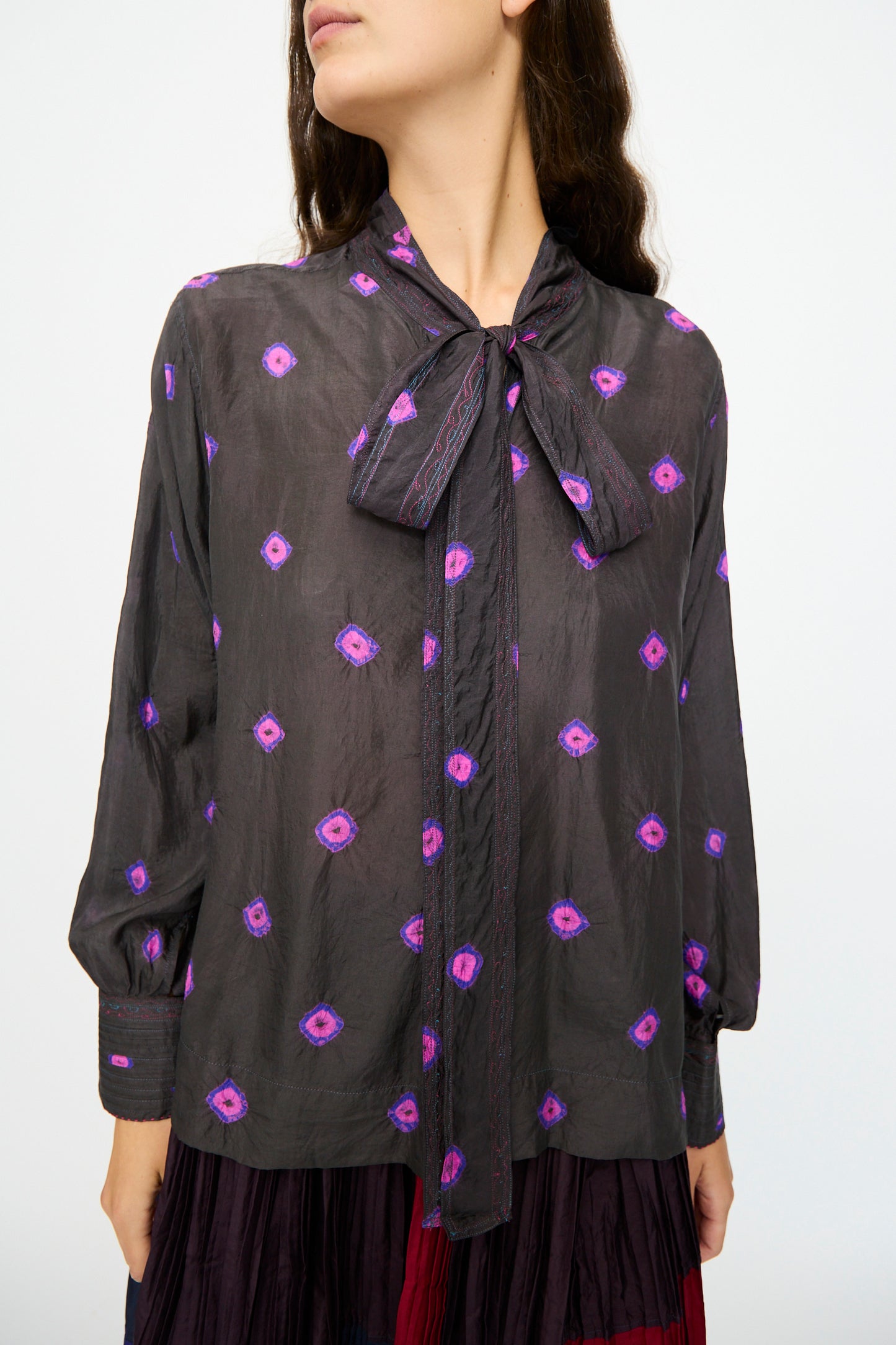 A woman wears the Injiri Silk Shirt in Purple, featuring sheer silk fabric, long sleeves, geometric patterns, and a tied neck bow.