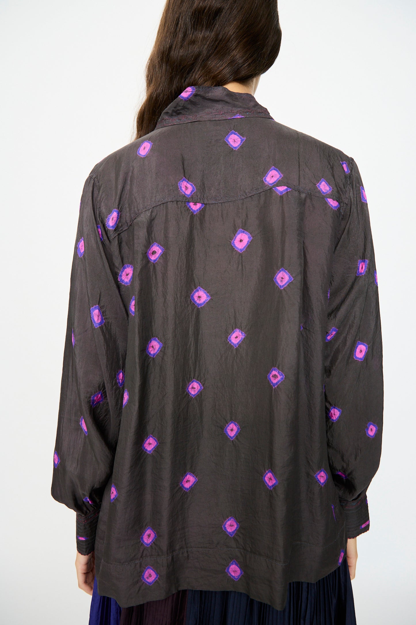 A person with long brown hair is seen from the back wearing the Injiri Silk Shirt in Purple—a loose-fitting, long sleeve blouse featuring a geometric pattern.