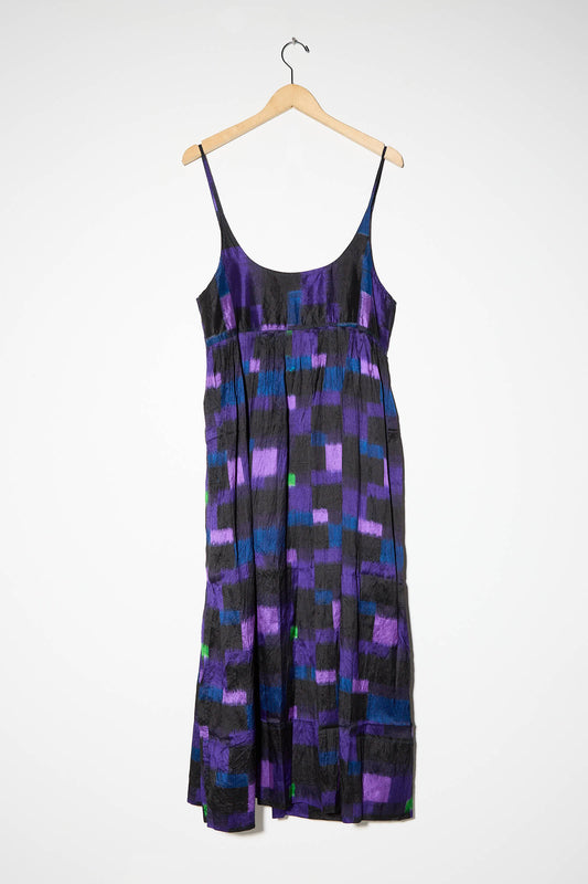 The Injiri Silk Slip Dress in Purple Multi, a sleeveless dress with adjustable thin straps and a multicolored abstract checker pattern featuring shades of purple, blue, and black, hangs on a wooden hanger against a plain white background.