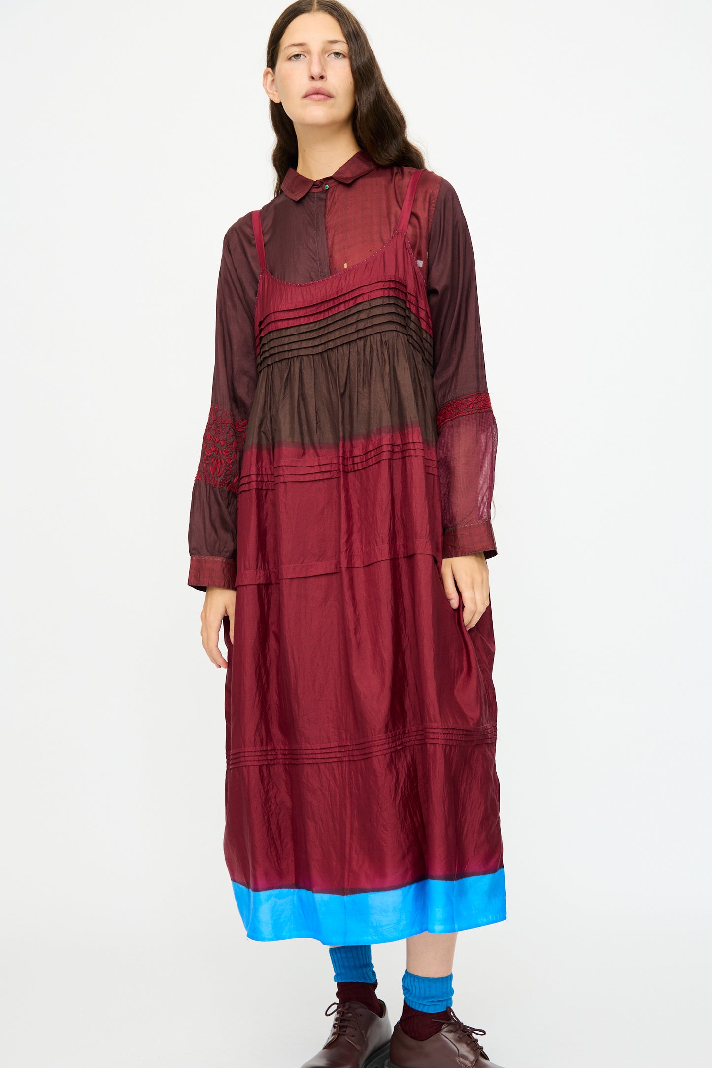 A person wears the Injiri Silk and Cotton Slip Dress in Red, featuring a long, layered design with a bright blue hem, paired with brown shoes and blue socks against a plain white background.
